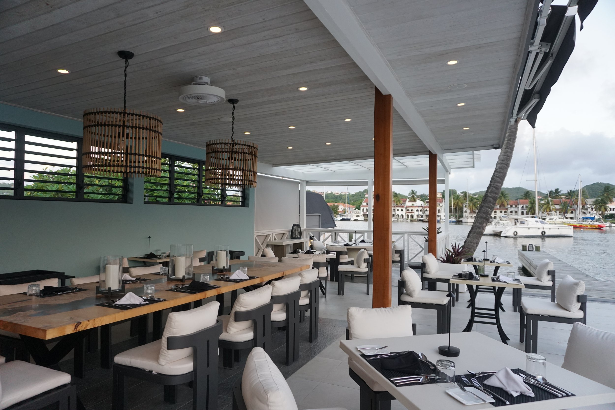 Empty restaurant with tables set for dining, overlooking a marina with boats and houses in the background, under a covered patio with pendant lights.