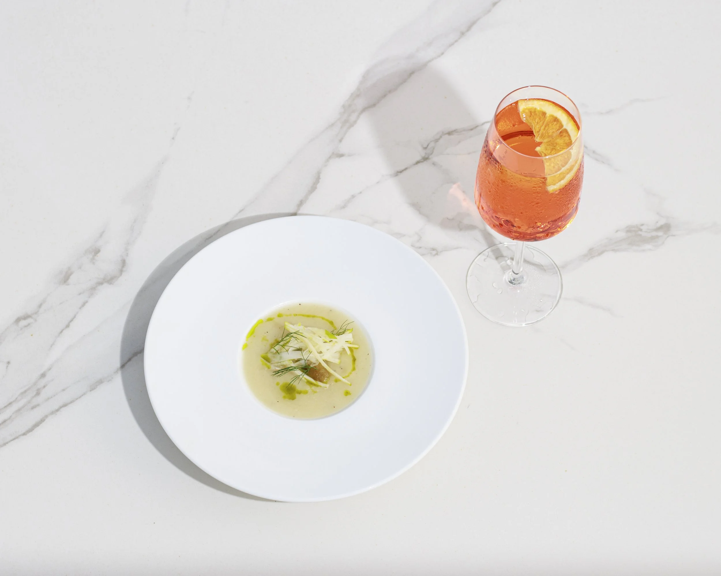 A white plate with soup garnished with herbs next to a glass of pink cocktail with a lemon slice on a white marble surface.