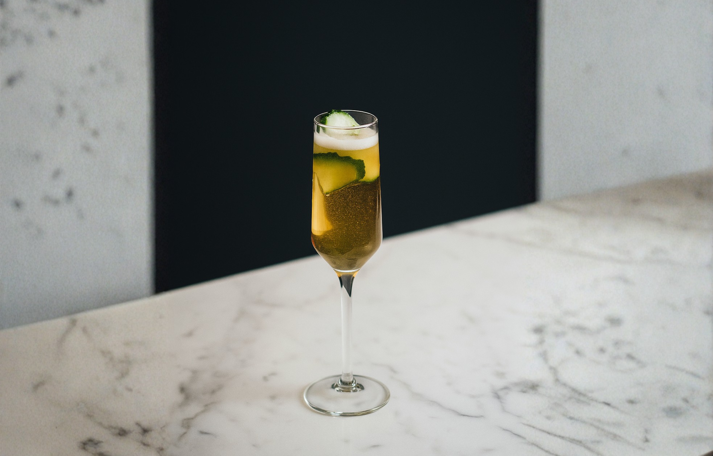 A tall glass of champagne with cucumber slices and a foam topping on a white marble surface.