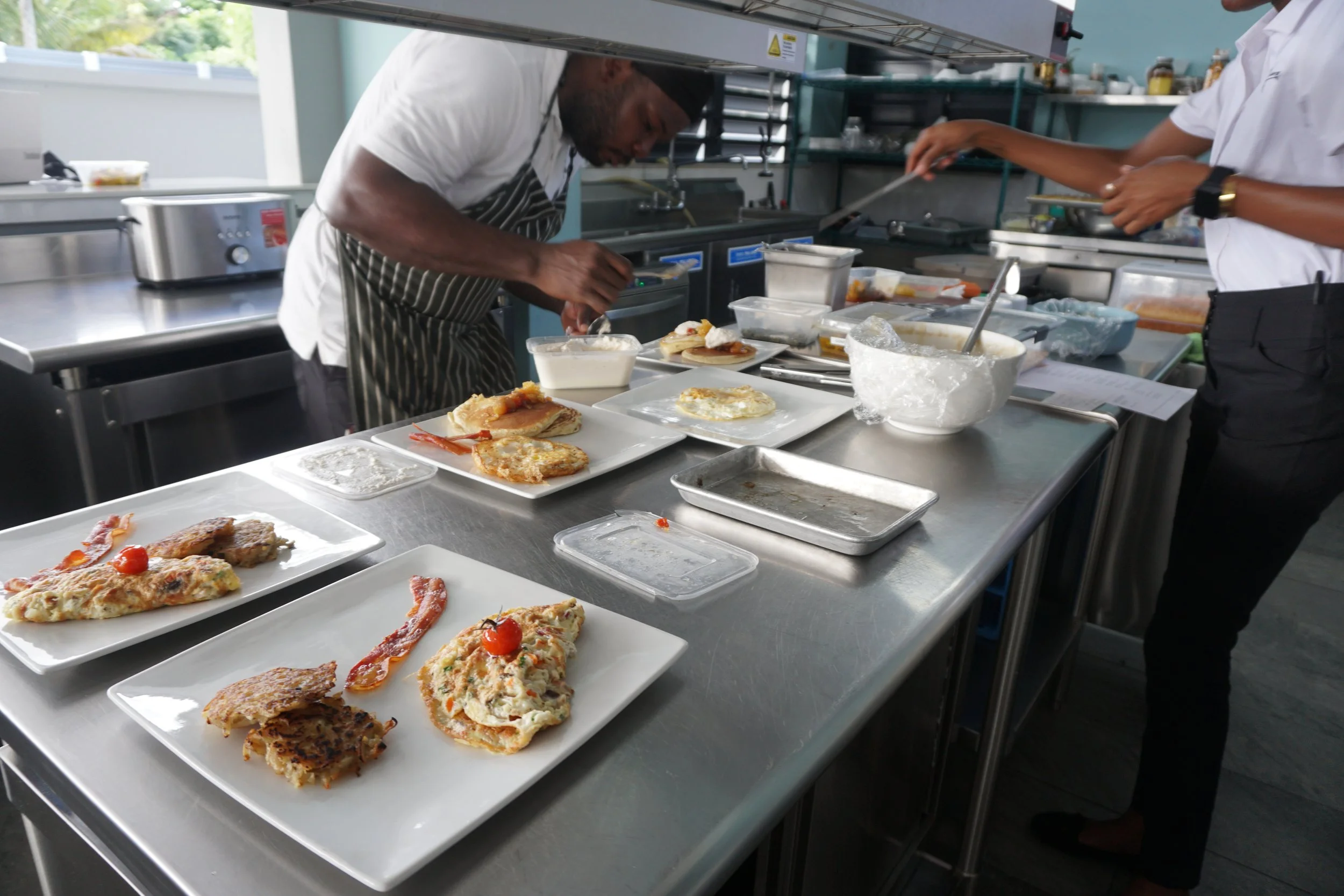 Two chefs preparing dishes with various plates of food on a stainless steel counter at The Harvest and Table Restaurant.