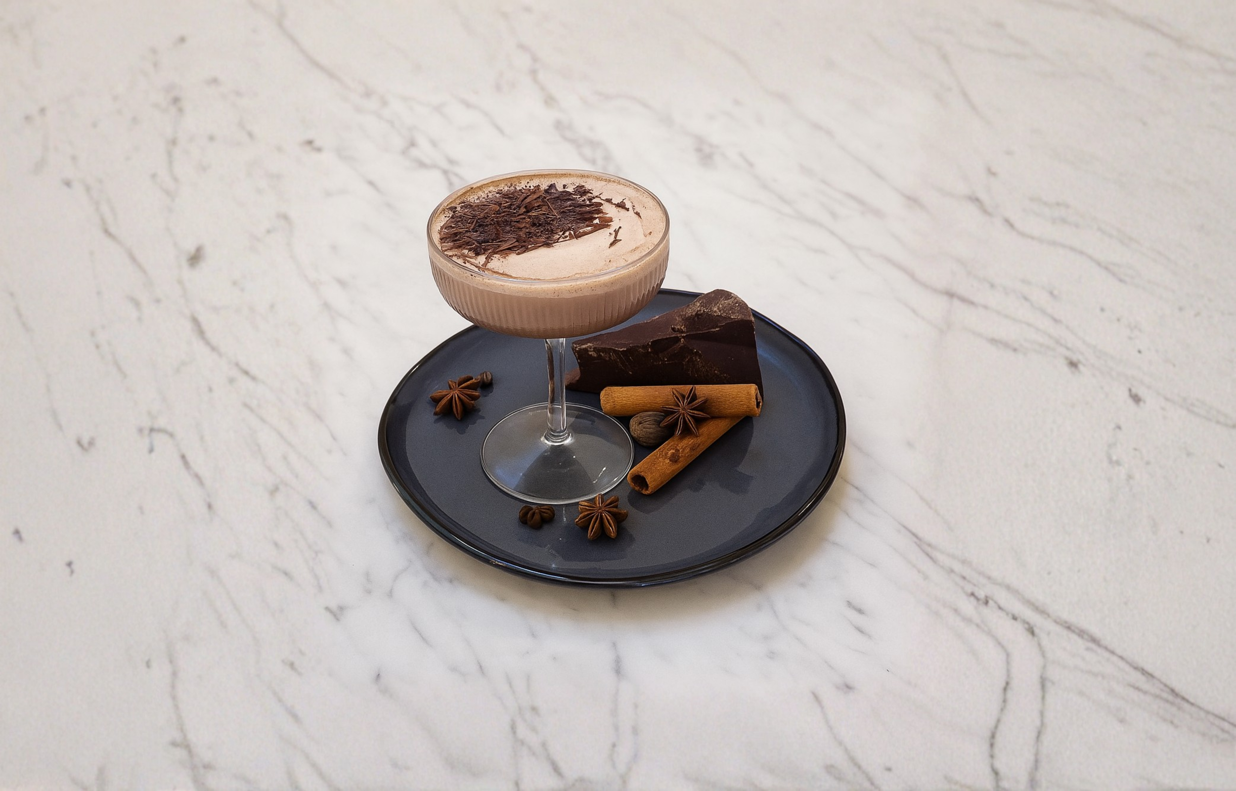 Chocolate cocktail garnished with chocolate shavings, served with chocolate pieces, cinnamon sticks, star anise, and coffee beans on a dark tray.