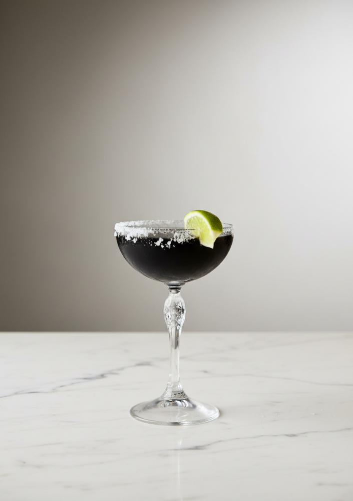 A black cocktail in a coupe glass with a salt rim and a lime wedge garnish, placed on a white marble surface.