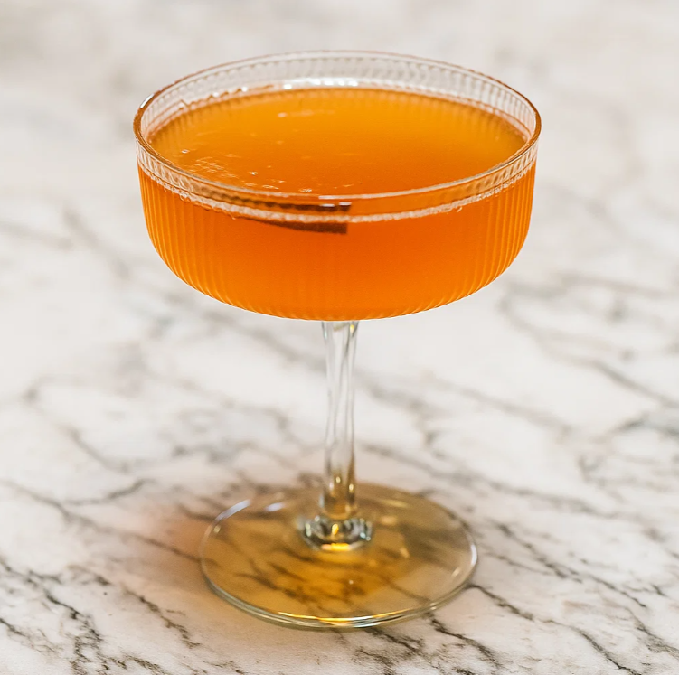 An orange cocktail in a coupe glass on a marble surface.