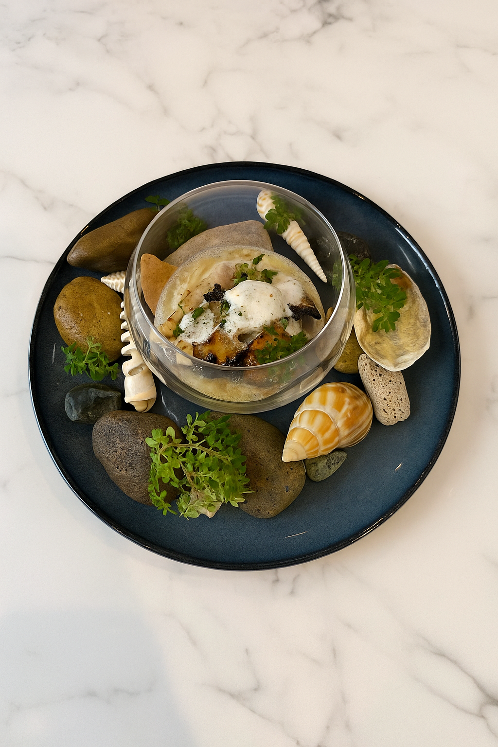 A black plate of assorted rocks, seashells, and green plant garnishes surrounding a glass bowl of gourmet food with foam, herbs, and roasted vegetables on a white marble surface.