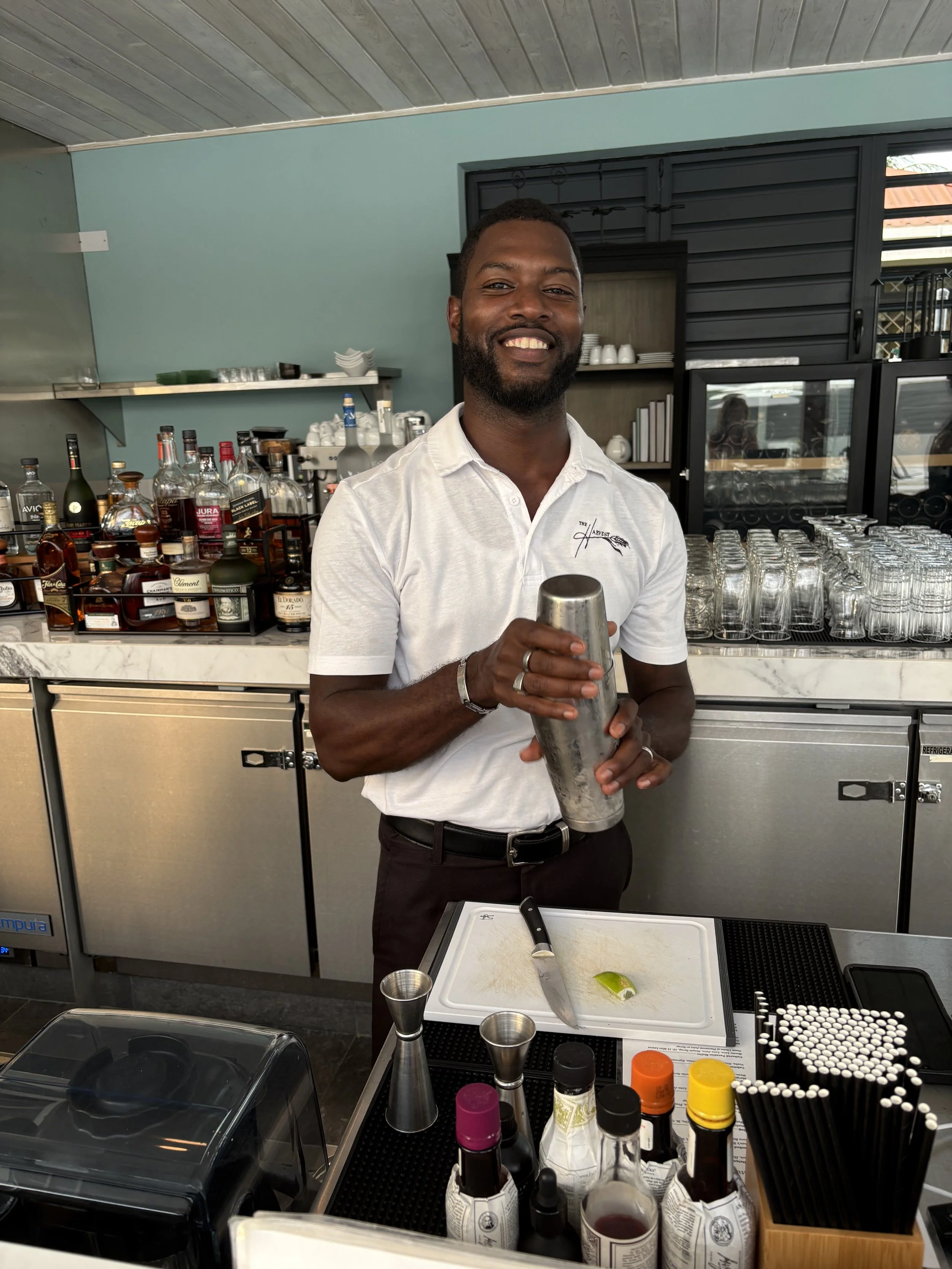 A smiling bartender with a beard, wearing a white polo shirt, stands behind a bar at The Harvest and Table Restaurant. 