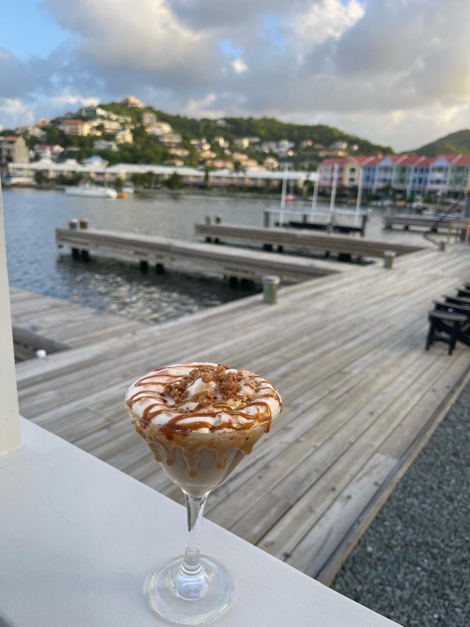 A dessert in a martini glass topped with whipped cream, caramel drizzle, and crumbled cookies, with a waterfront view of docked boats, a wooden deck, and hillside houses in the background under a partly cloudy sky.