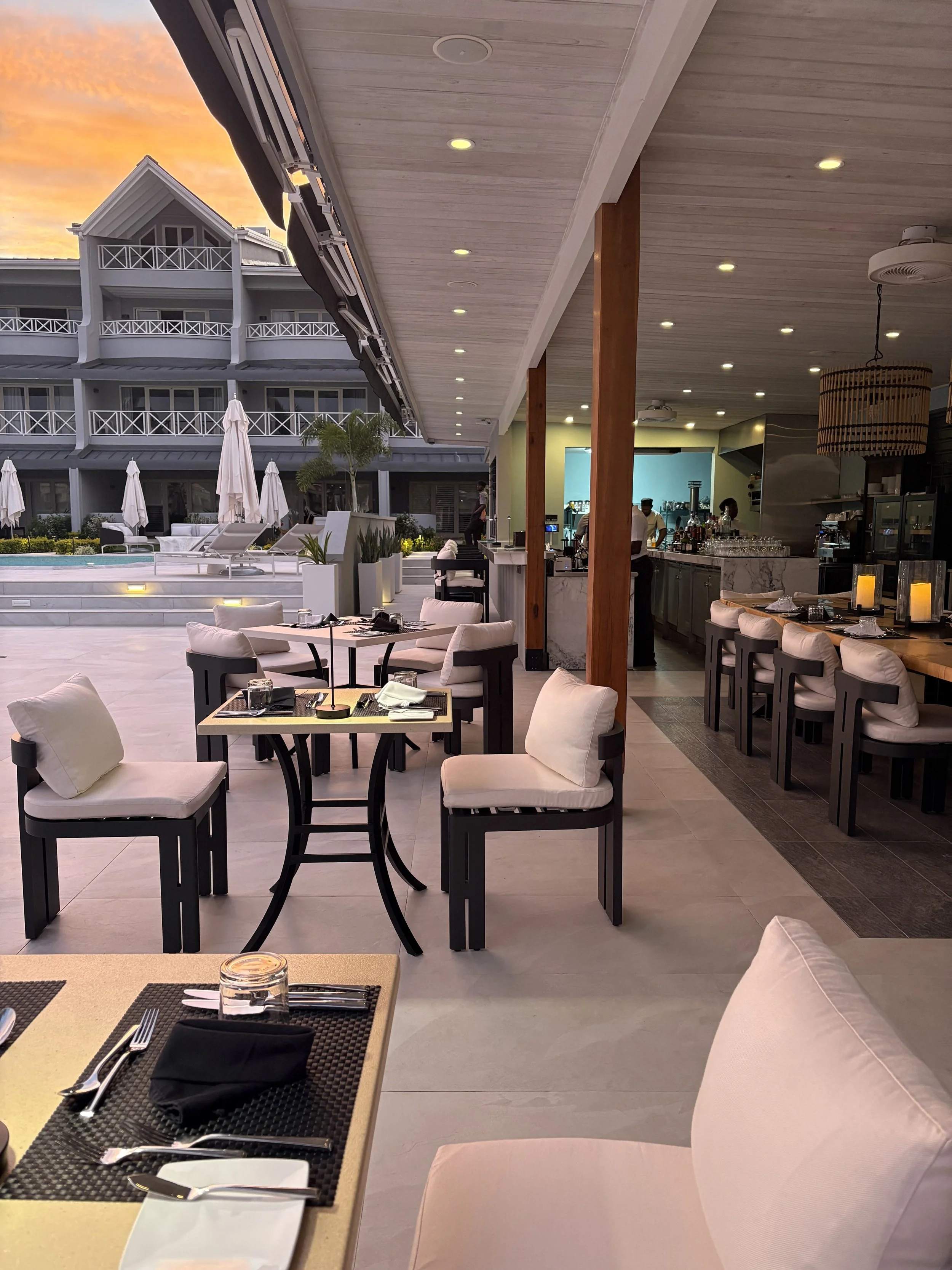 Outdoor dining area at a restaurant with a pool and hotel building in the background during sunset. The dining area has tables with cushioned chairs, set with black napkins, utensils, and glassware.
