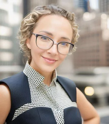 A woman with curly blonde hair wearing glasses, a polka-dot blouse, and a navy blazer stands outdoors in an urban environment with buildings in the background.