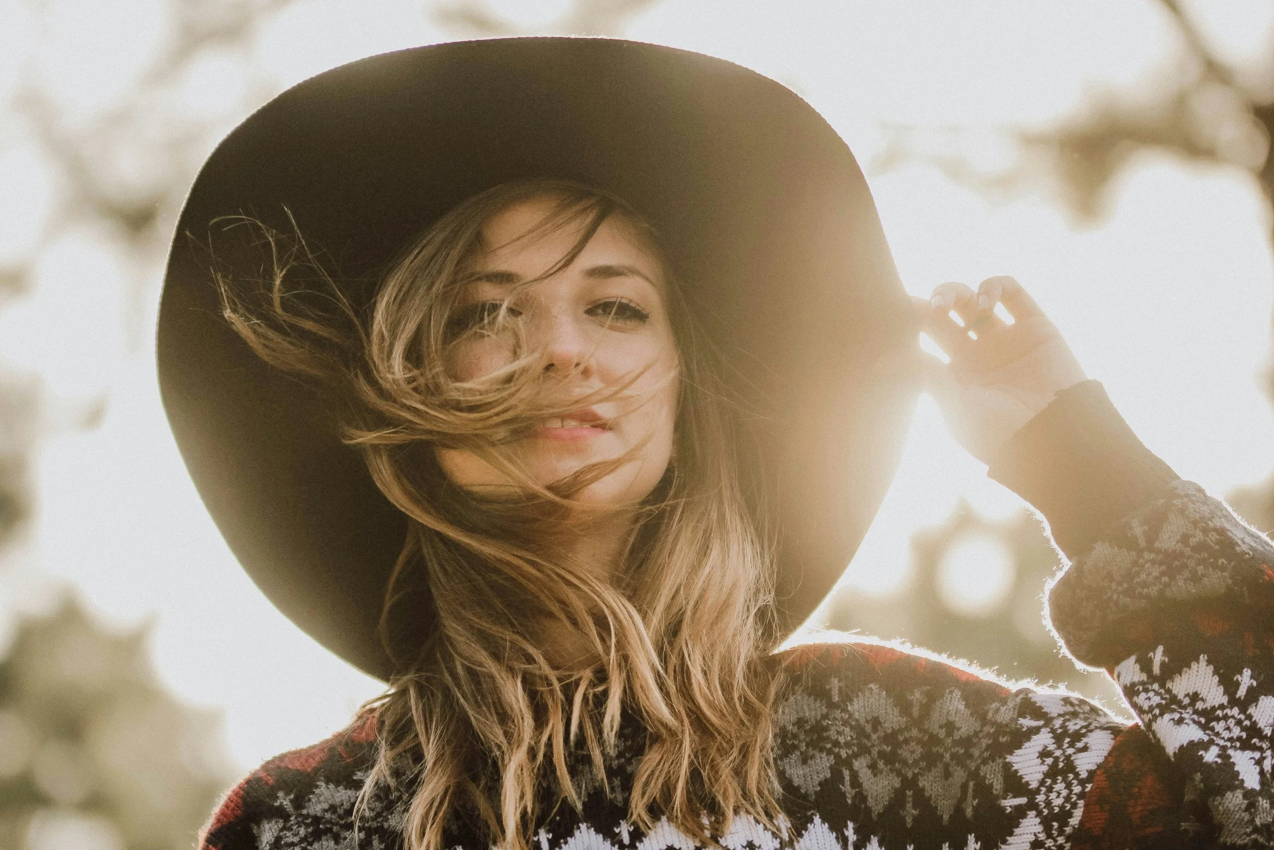 Young woman outdoors wearing a large black hat and patterned sweater, smiling and holding the brim of her hat, with sunlight behind her.