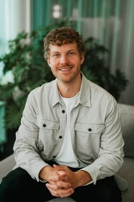 Ung man med krulligt hår och vit jacka som ler, sittandes framför gröna växter.