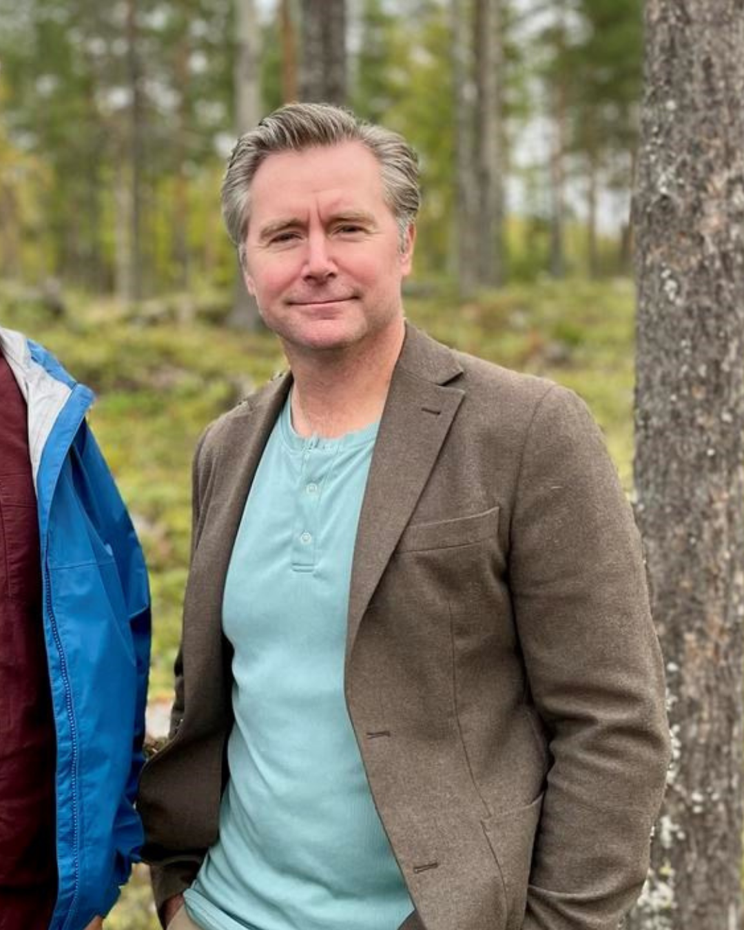 En man med grått hår och lätt skägg, iklädd en ljusblå tröja och brunt kavaj, står i en skog med träd i bakgrunden.