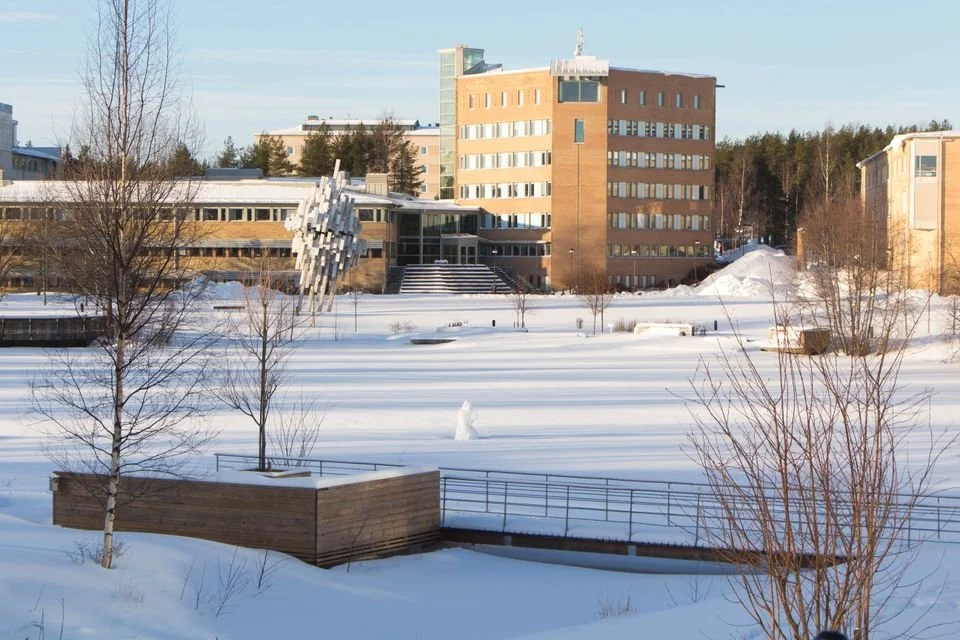 Snötäckta byggnader och träd på en vintrig dag, med ett vattenområde täckt av snö och en blå himmel i bakgrunden.