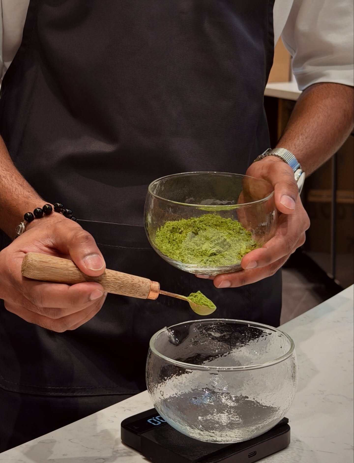 Matcha Bar at Centro Singapore, Shaw theatres, Lido cinema. Person in dark apron and white shirt preparing matcha tea, using a bamboo scoop to transfer green powder into a glass bowl, on a kitchen counter with a scale and clear bowl.