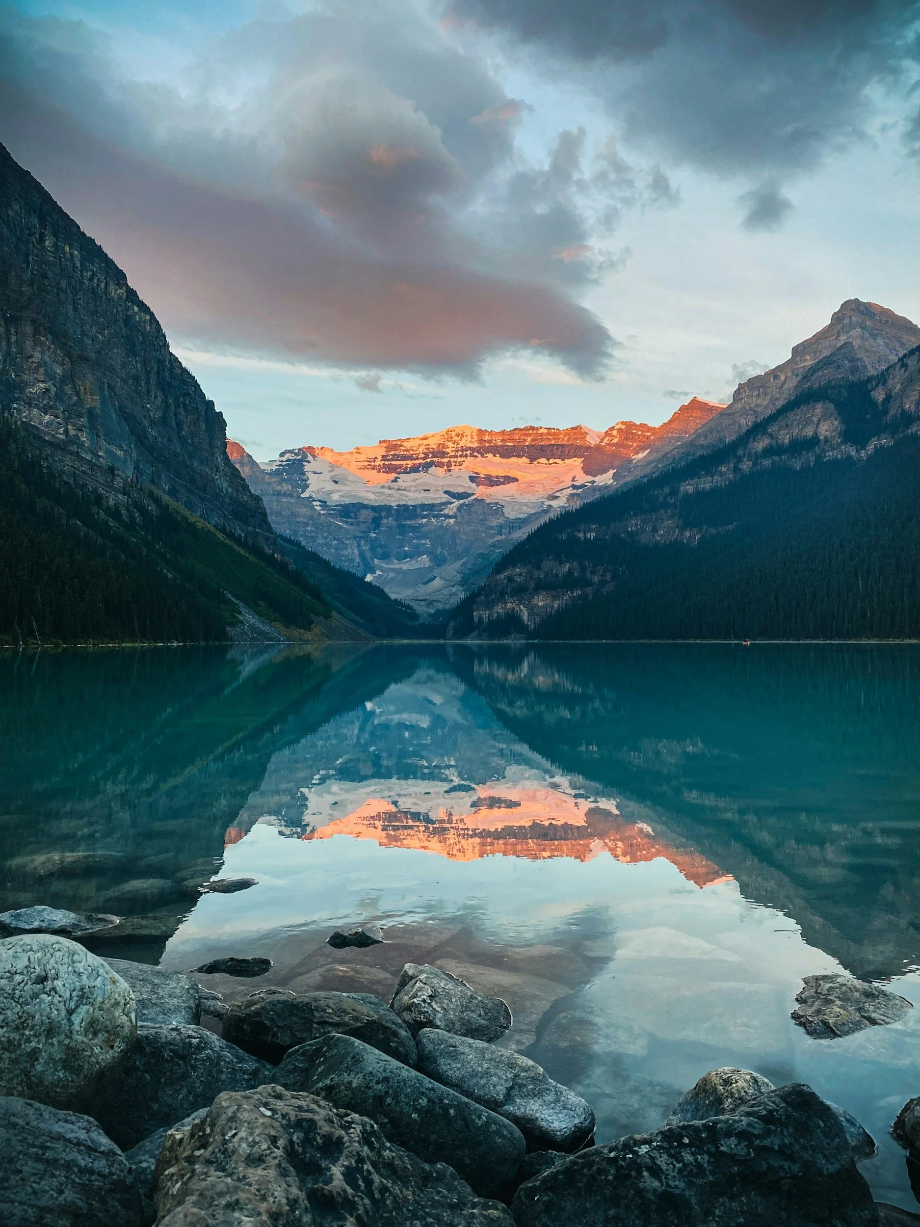 Scenic mountain lake reflection with rocky shoreline and snow-capped mountains under a cloudy sky at sunset.
