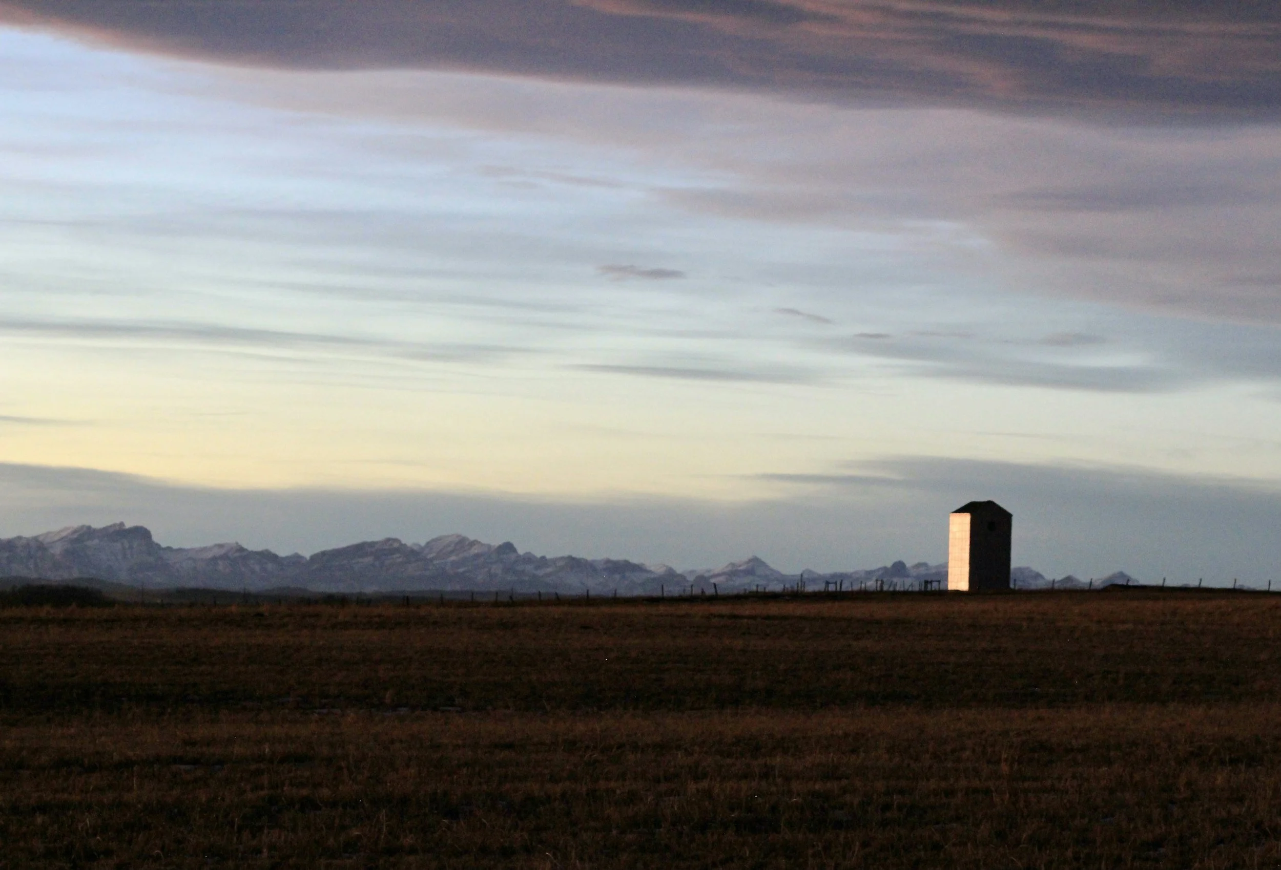 Open landscape with flat grassy field, distant snow-capped mountains, and a tall, narrow stone tower on the right side, under a cloudy sky during sunset or sunrise.