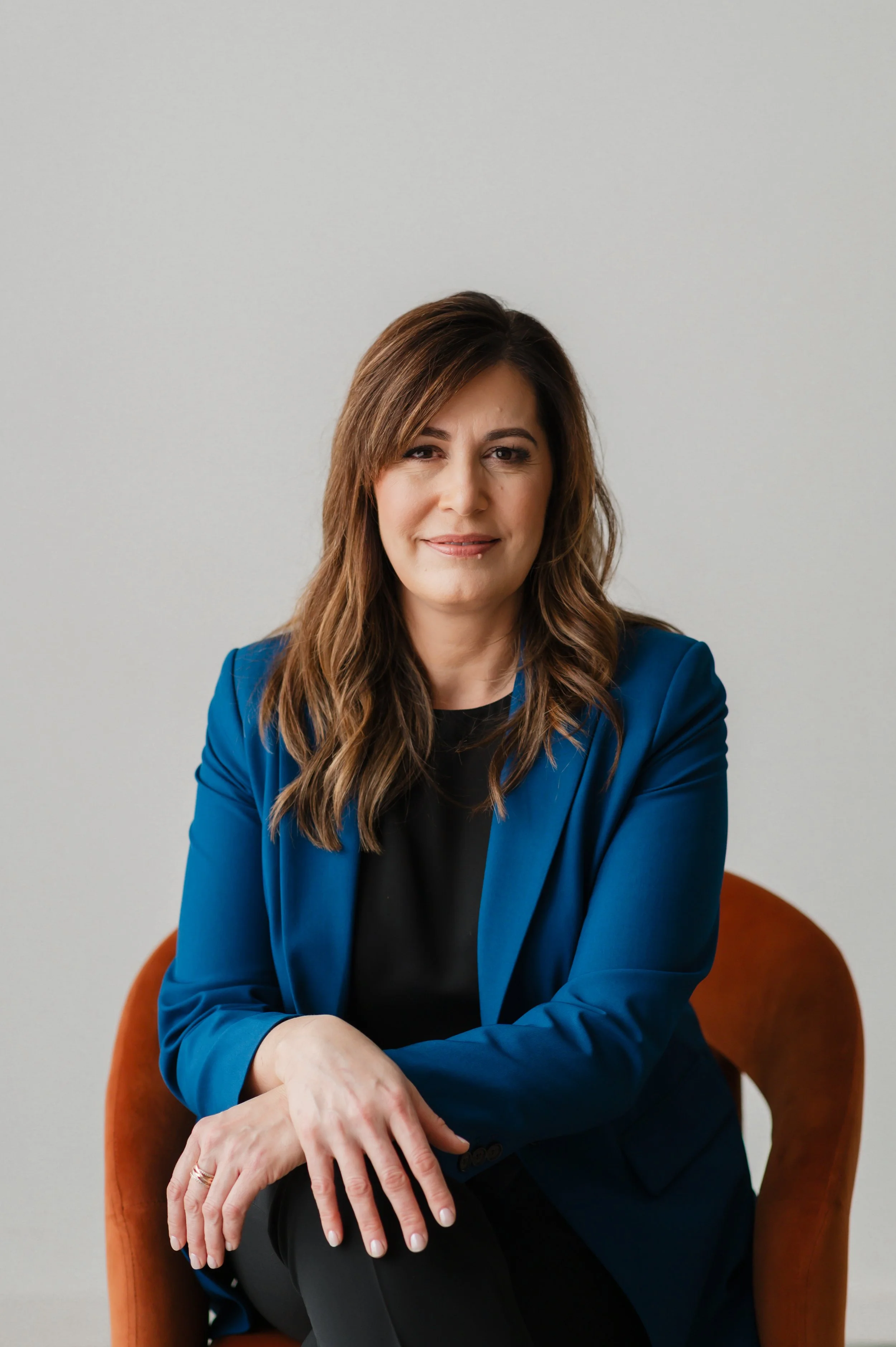 A woman with shoulder-length brown hair wearing a blue blazer and black top, sitting on an orange chair against a plain light-colored wall.