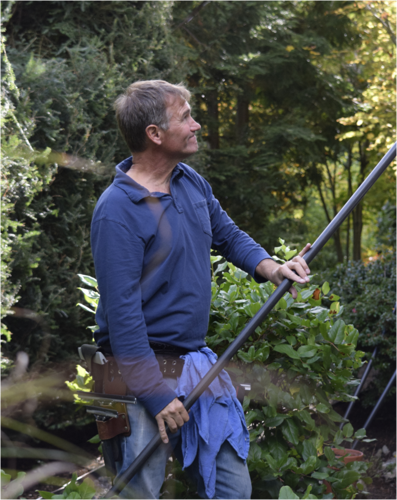 Man with a tool belt using a pole with a squeegee on the end in a garden with lush greenery and trees
