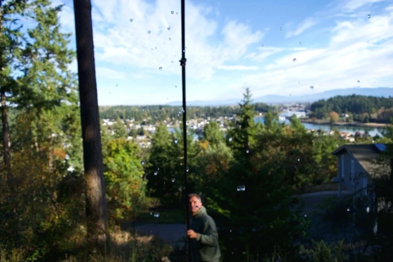 A man standing outdoors near a tall pole with water droplets on the camera lens, with trees and a town visible in the background.
