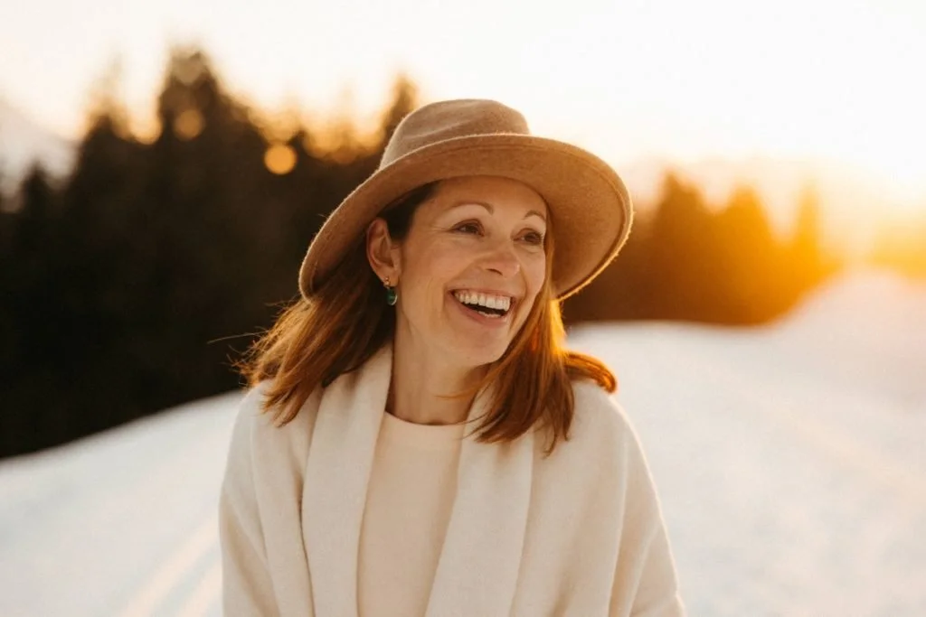 A woman smiling outdoors at sunset wearing a beige hat and a cream-colored coat.