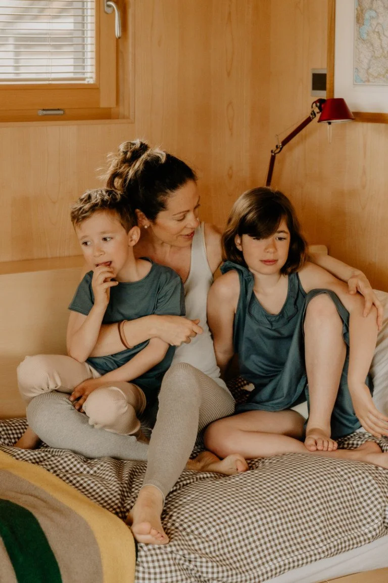 A woman and two children sitting on a bed in a wood-paneled room, with a window and a map on the wall in the background.