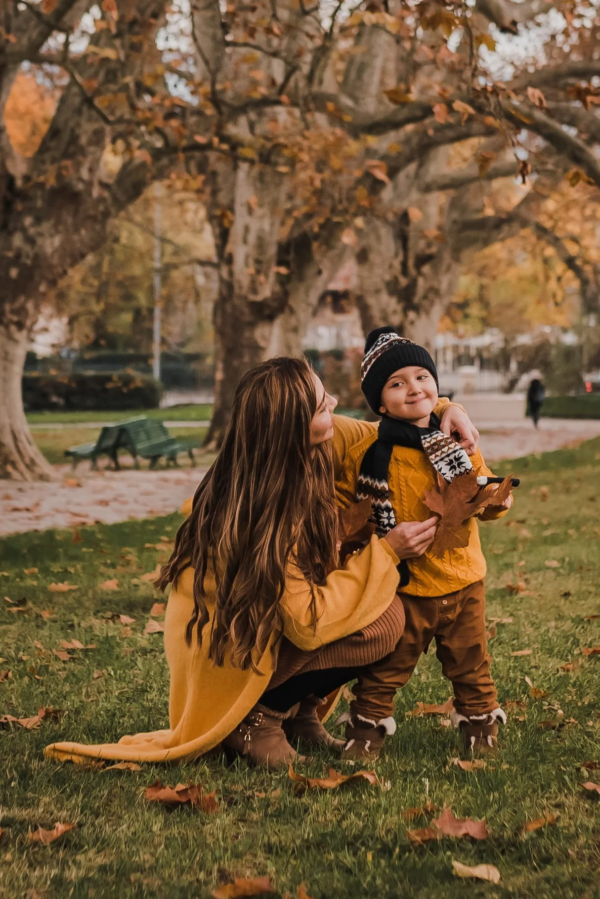 A woman crouching down and smiling at a young boy in a park during autumn, both holding large fallen leaves. The woman has long brown hair and is wearing a yellow coat, and the boy is dressed in a yellow jacket, brown pants, a black beanie, and a patterned scarf. The background features trees with orange and brown leaves, park benches, and a person walking in the distance.