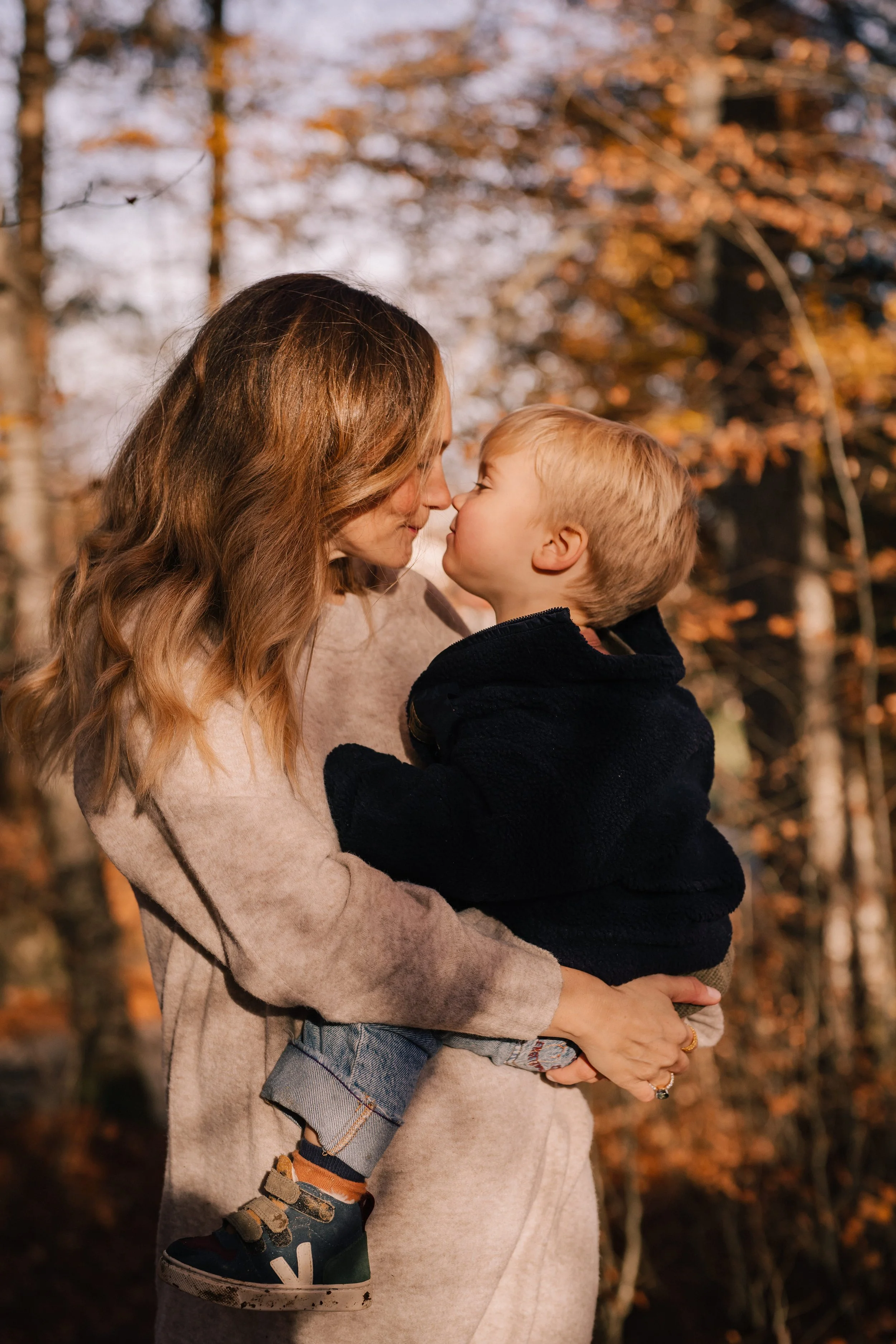 A woman holding a young boy in an outdoor setting with fall foliage in the background, sunlight filtering through trees.