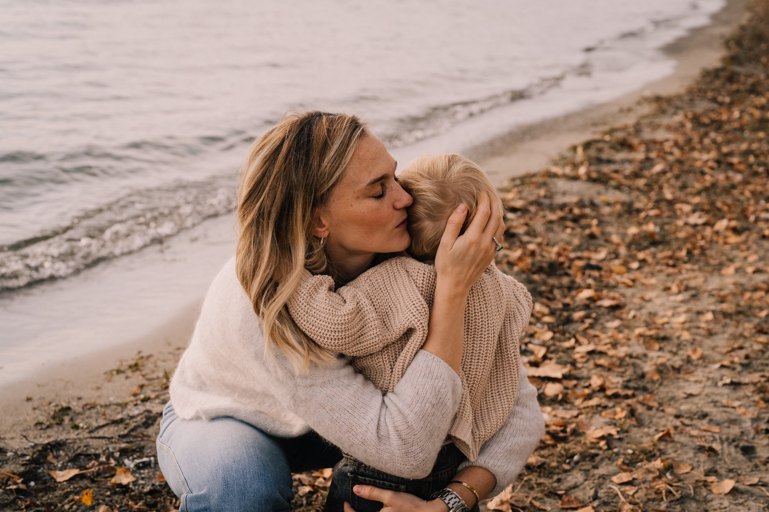 A woman embraces and kisses a child on a beach with fallen leaves, near the water's edge.
