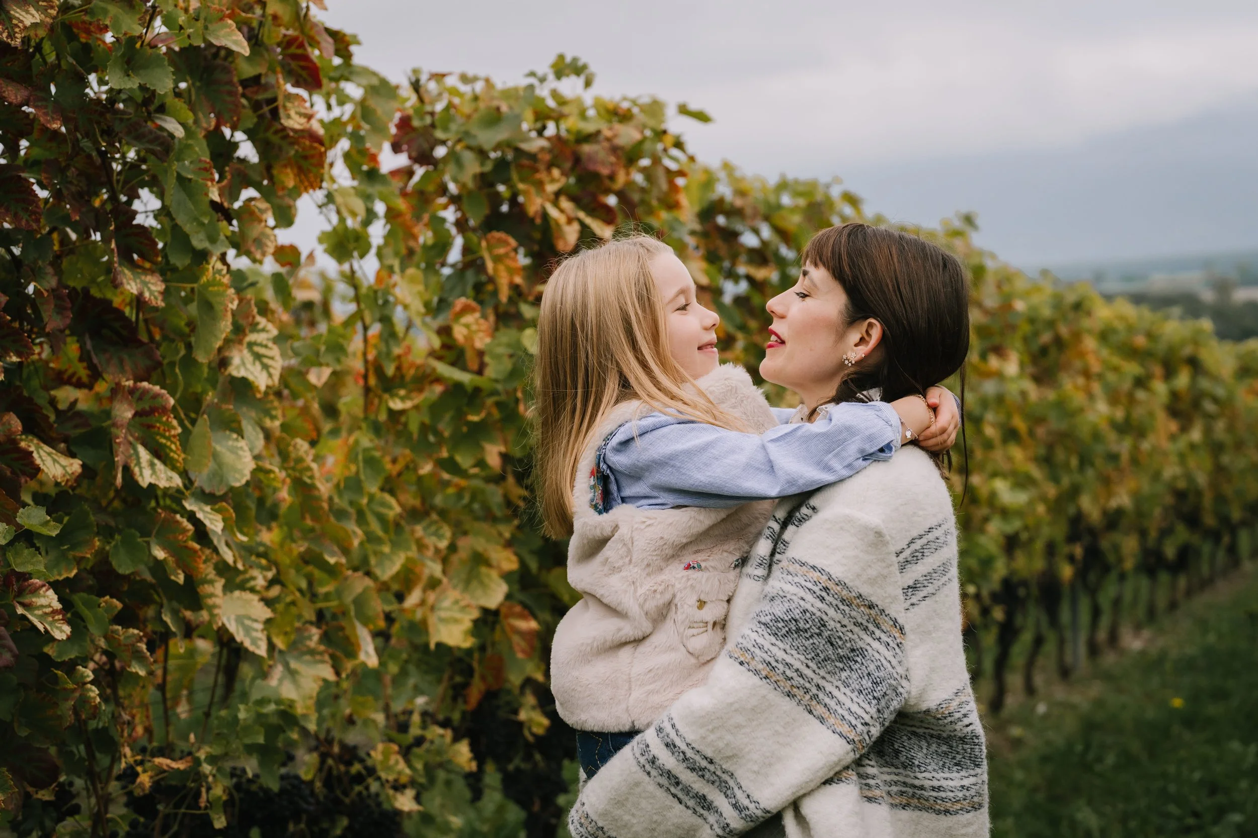 A woman holding a young girl in a vineyard with green and yellow leaves.