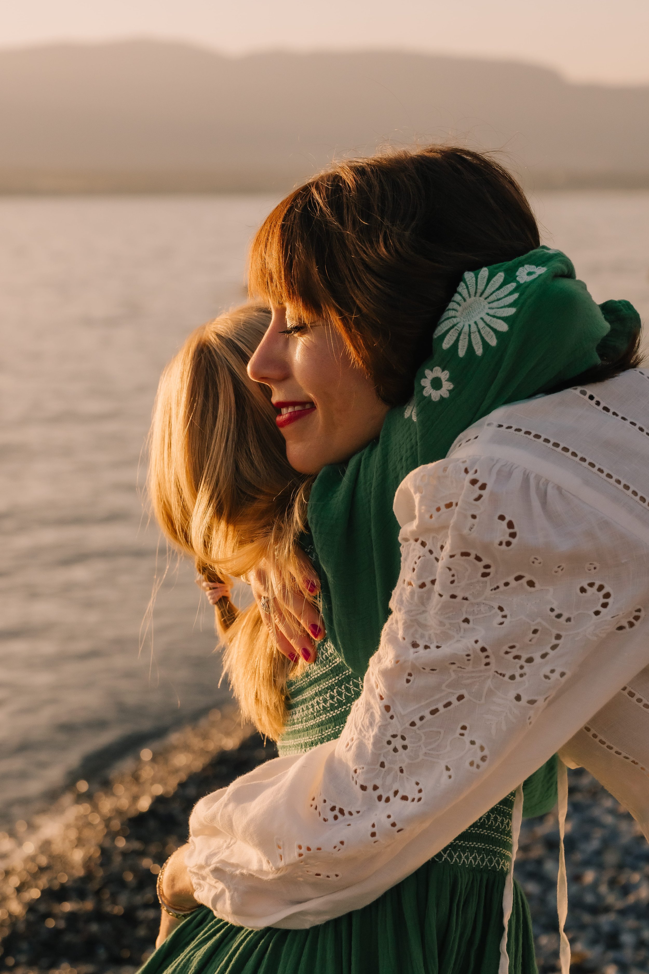 A woman hugging a young girl with long blonde hair on a beach during sunset.