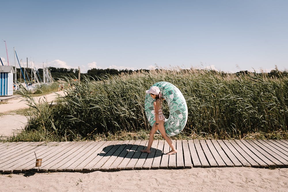 A girl walking on a wooden pathway at the beach carrying a large inflatable inner tube, wearing a sun hat and swimsuit, with sailboats and grassy dunes in the background under a clear sky.