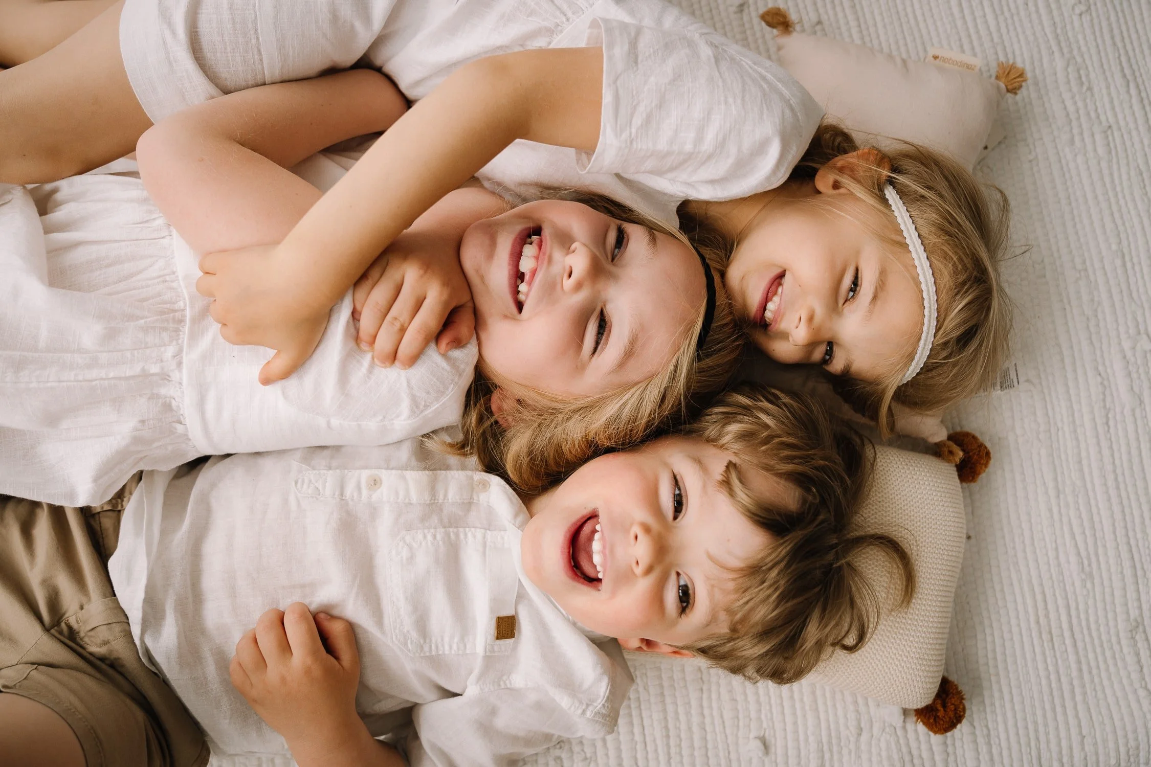 Three children lying on a bed, smiling and hugging each other.