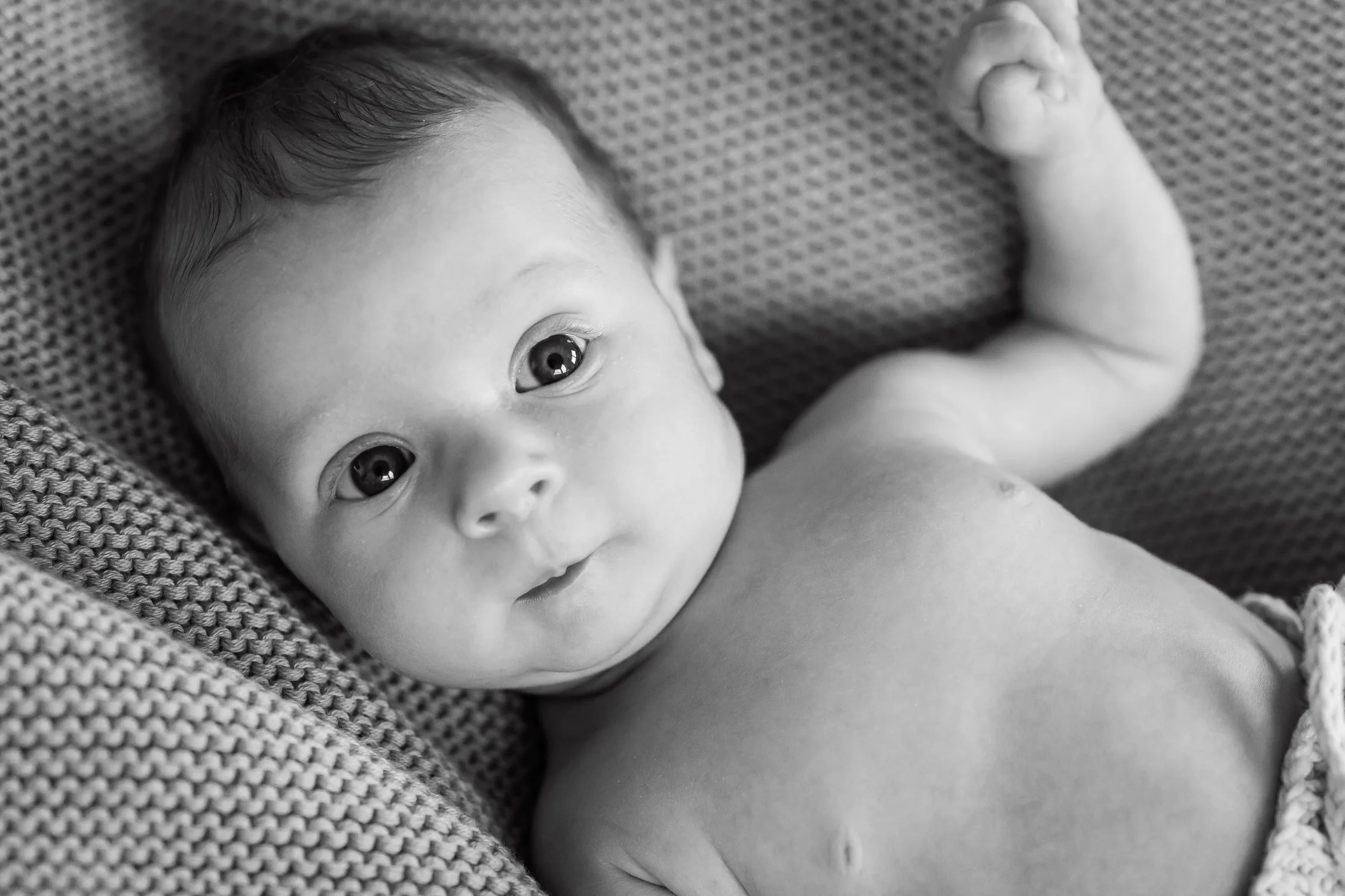 Black and white photo of a baby lying on a textured blanket, looking directly at the camera with wide eyes and a slight smile.
