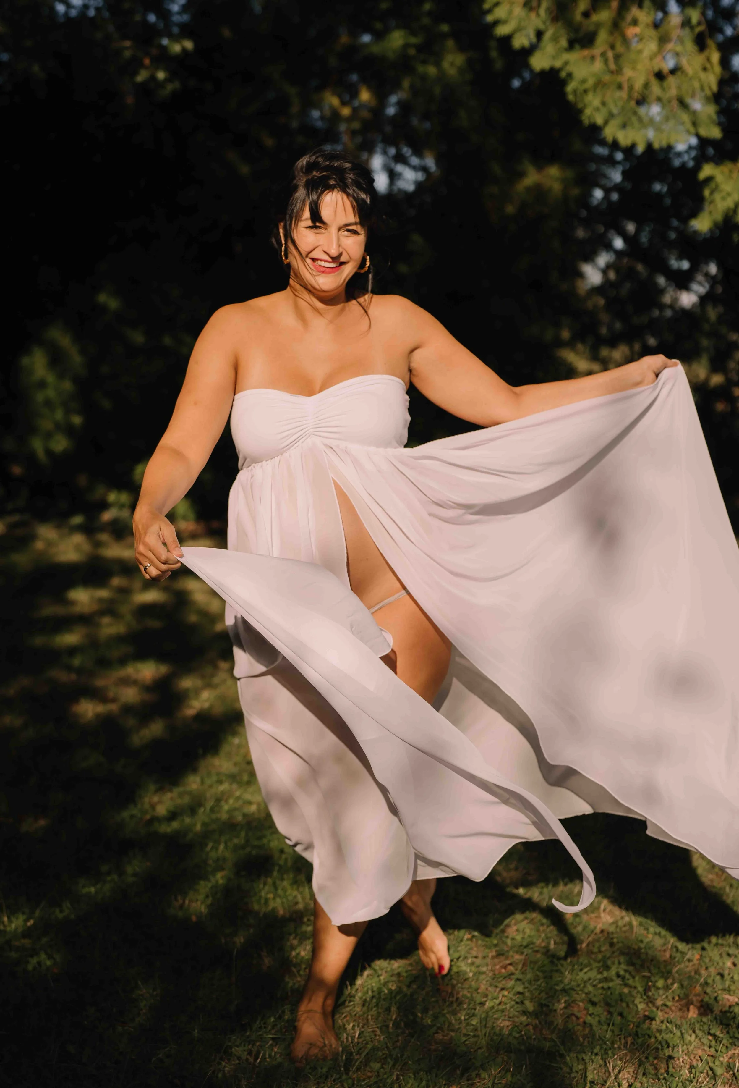 A smiling woman dressed in a white strapless dress is outdoors on grass with trees in the background, lifting her dress to reveal her leg and underwear.