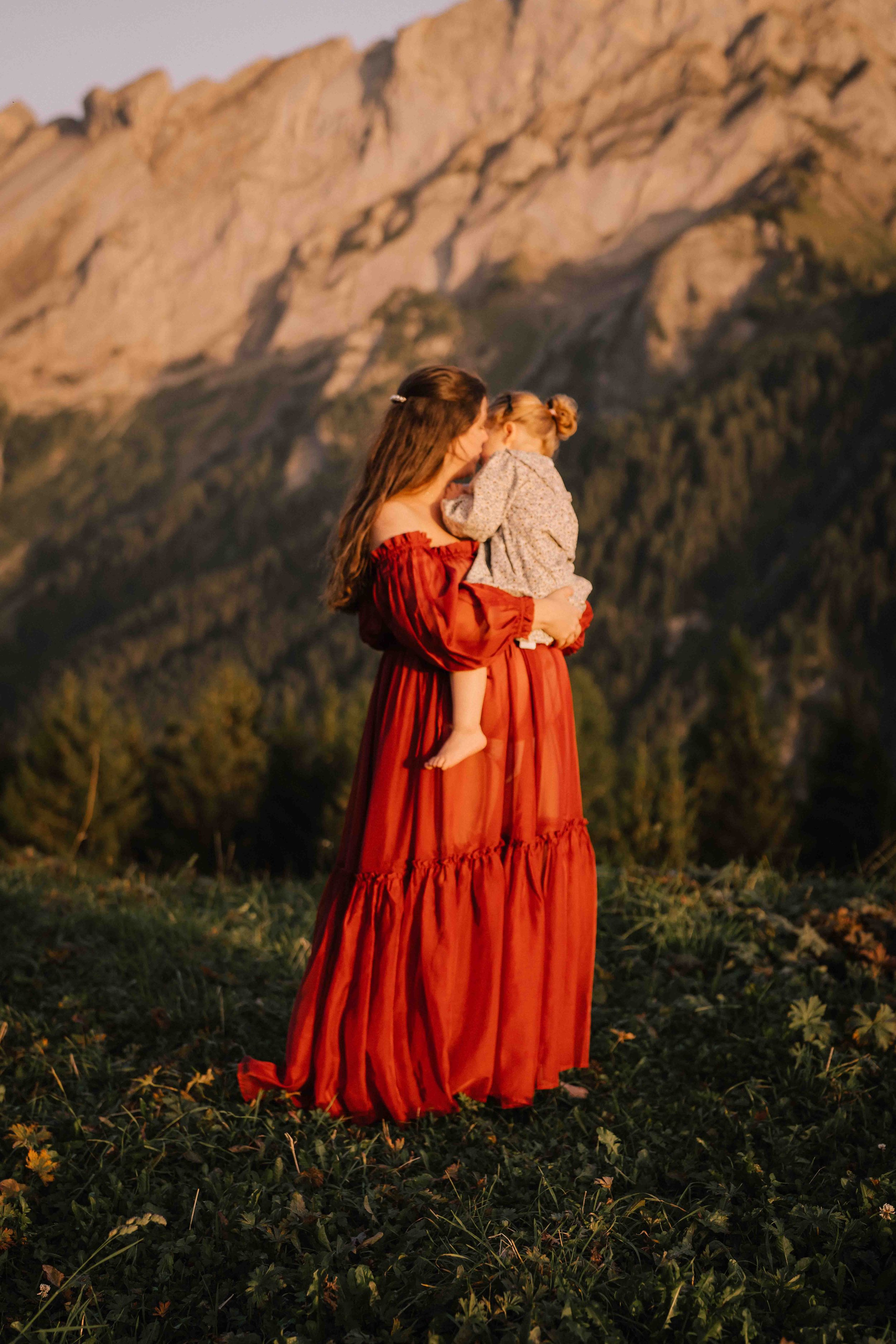 A woman in a long red dress holding a child, with a mountain landscape in the background during sunset.
