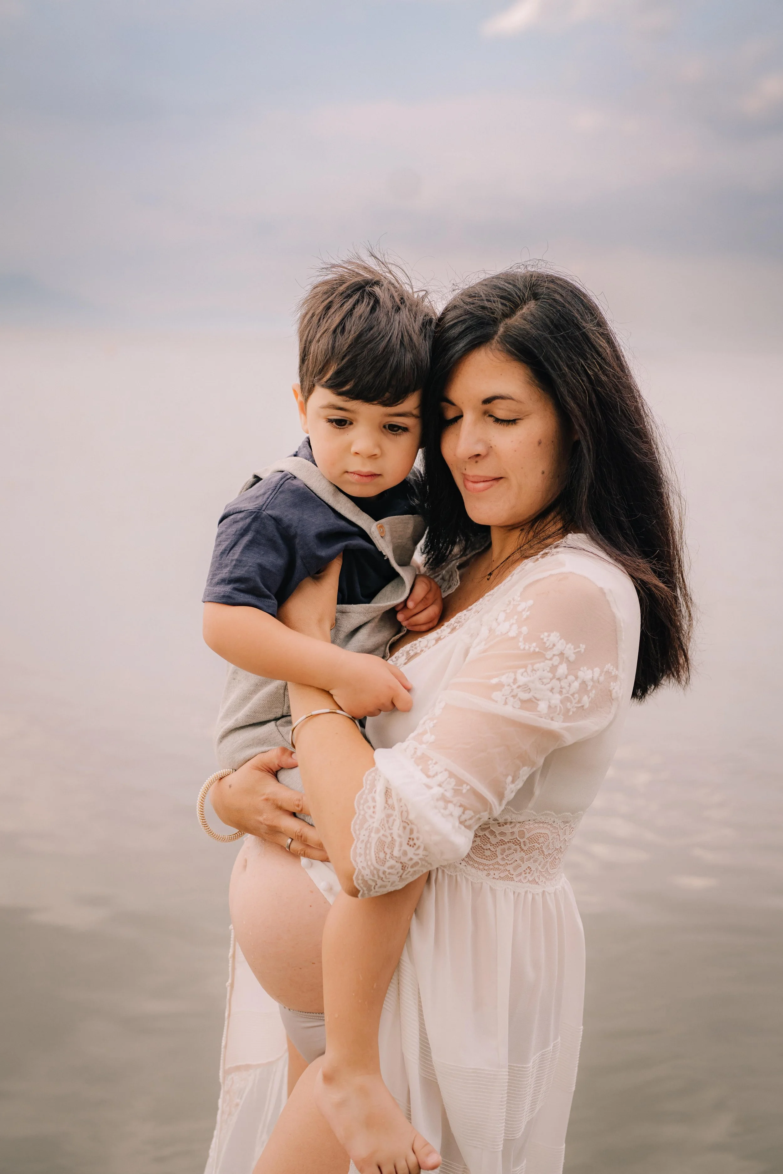 A woman holding a young boy at the beach during daytime with calm water and cloudy sky in the background.