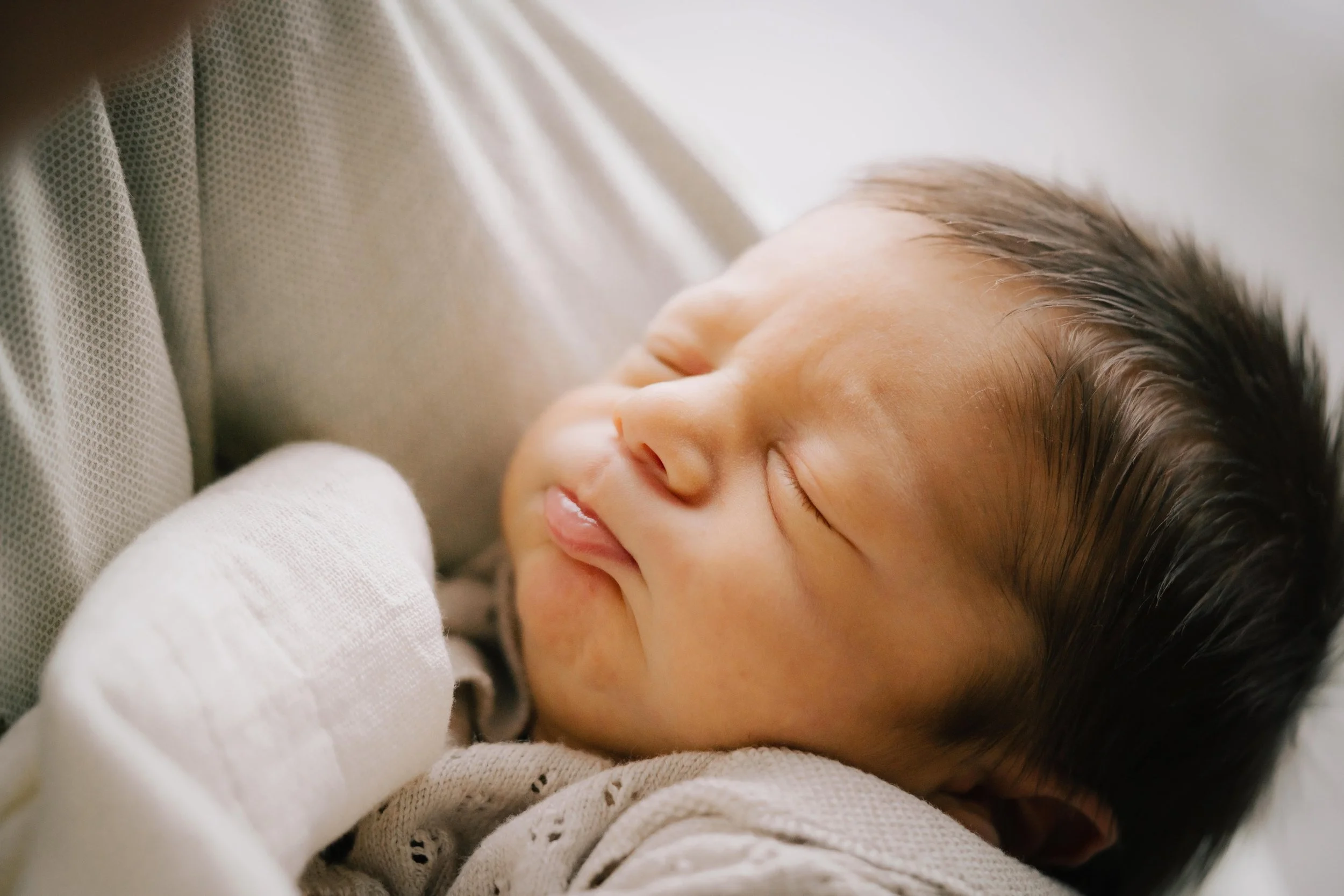 Close-up of a sleeping baby with closed eyes and a peaceful expression, dressed in light-colored clothing, resting against a soft surface.