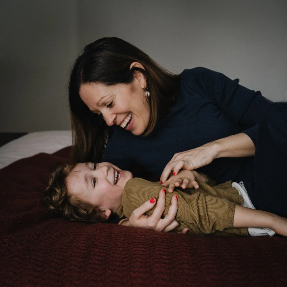 A woman and a young girl are playing or cuddling on a bed, smiling and looking at each other.