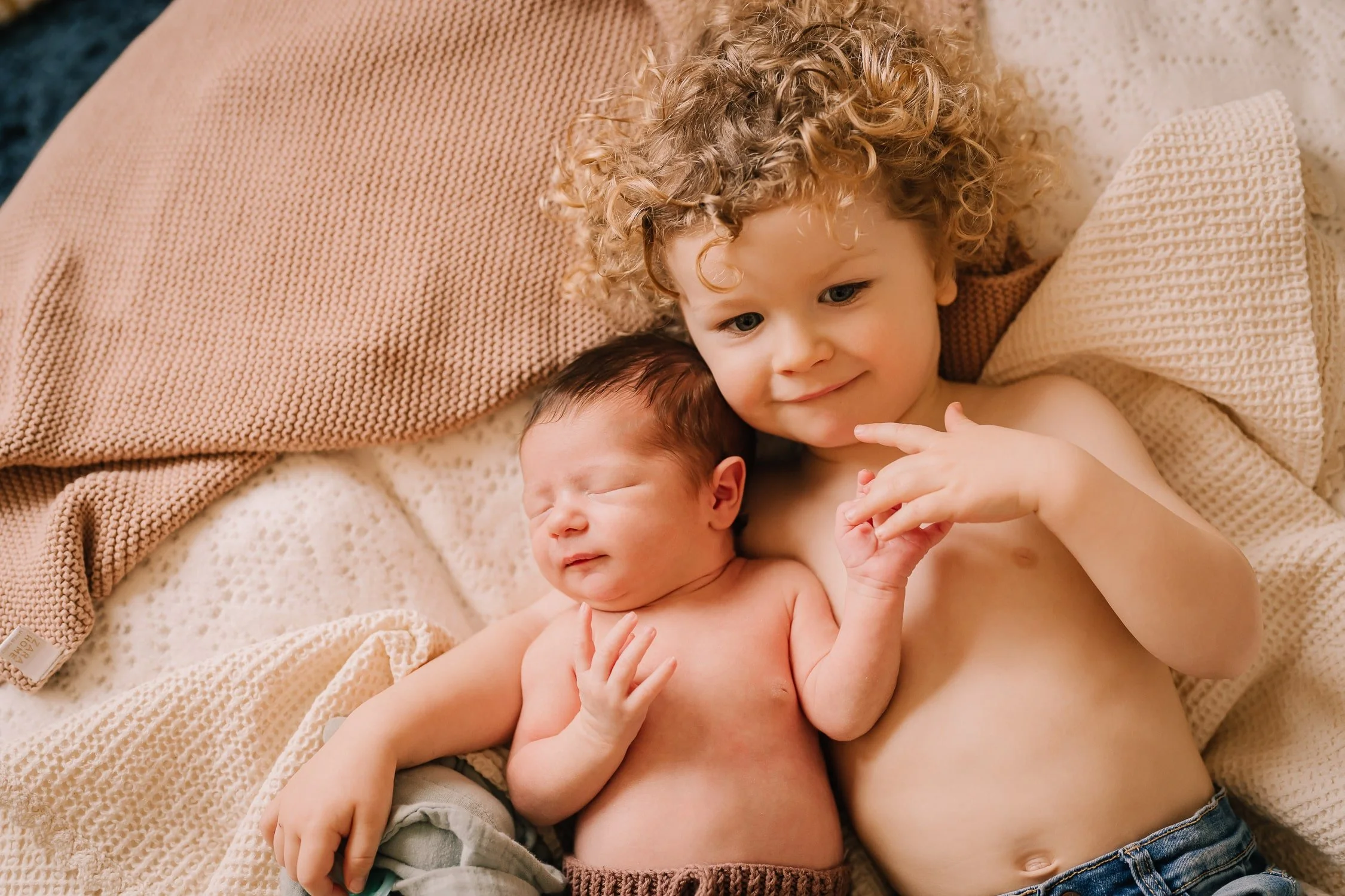 Two young children, a toddler with curly blonde hair and a newborn with dark hair, lying on a bed covered with beige blankets and pillows, sharing a tender moment.