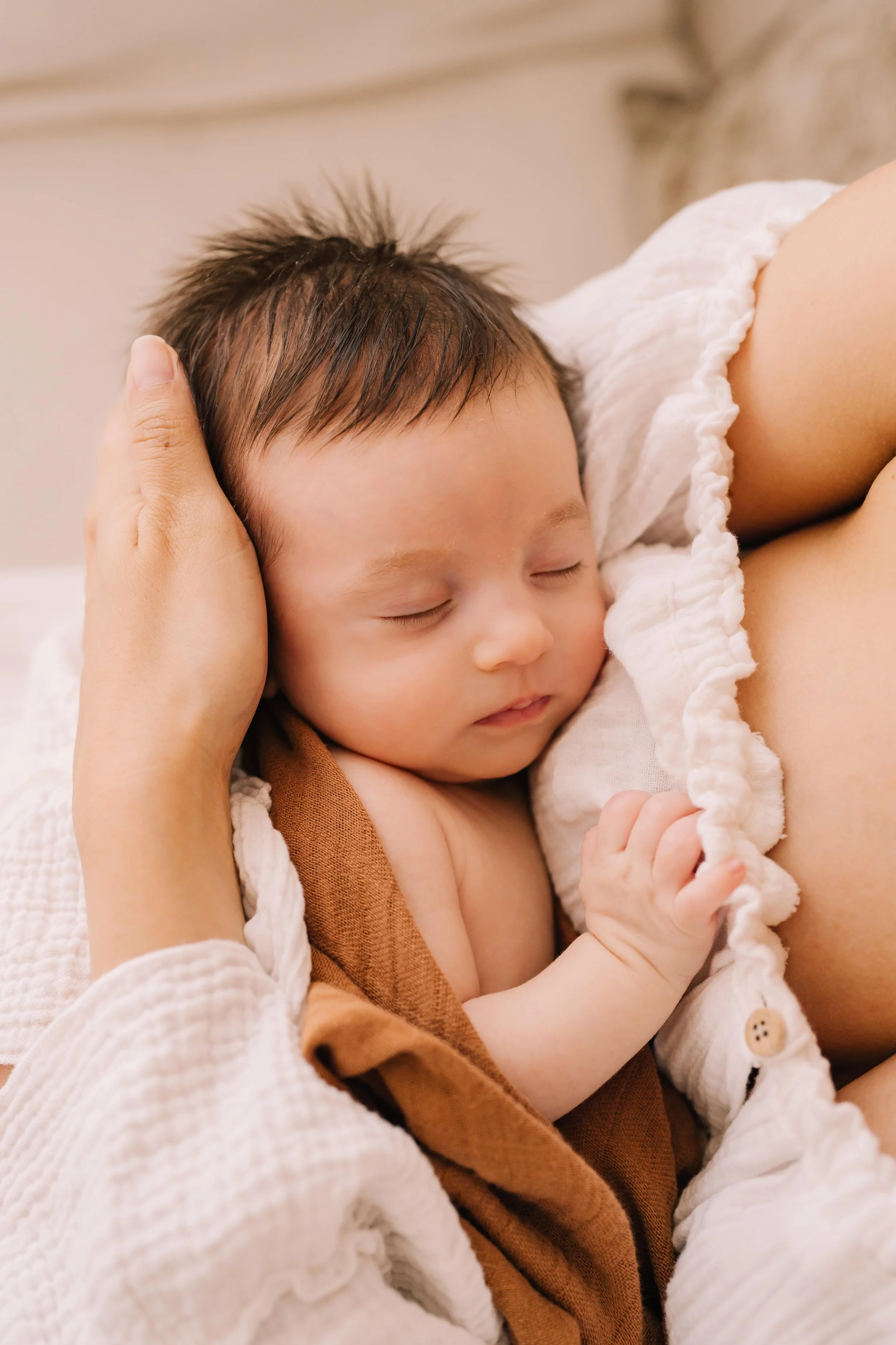 Close-up of a sleeping baby resting on a woman, who is gently holding the baby's head and breastfeeding. The baby has dark hair and is covered with a white, ruffled cloth. The scene takes place in a cozy, softly-lit setting.