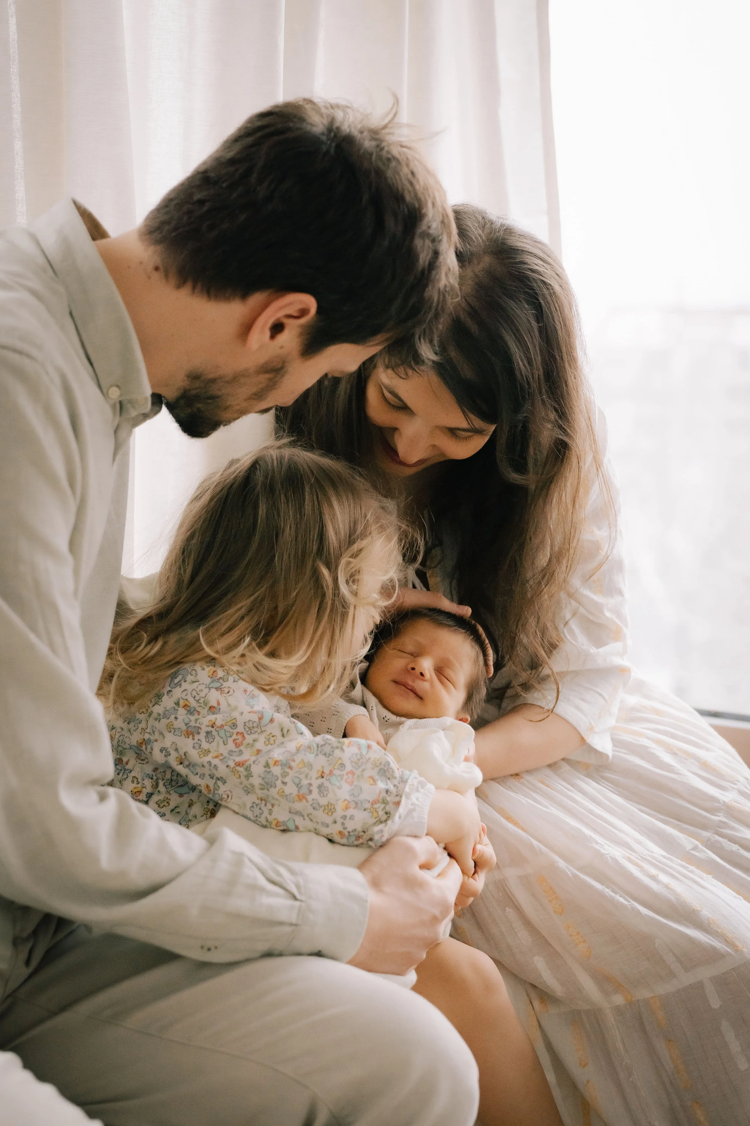 Family of four, including a man, woman, young girl, and baby, sitting on a bed by sunlight, sharing a joyful moment.