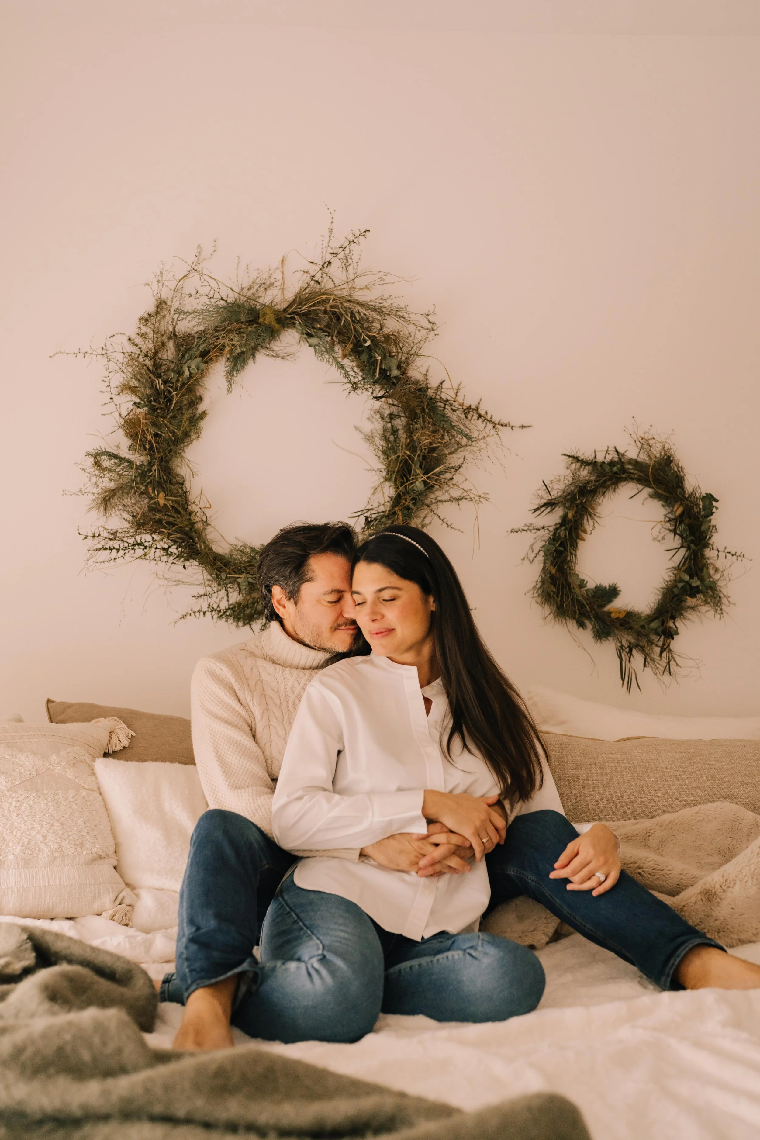A loving couple sitting on a cozy bed, embracing each other with closed eyes, surrounded by warm blankets and pillows. Behind them are three decorative circular wreaths made of twigs and greenery on a plain wall.