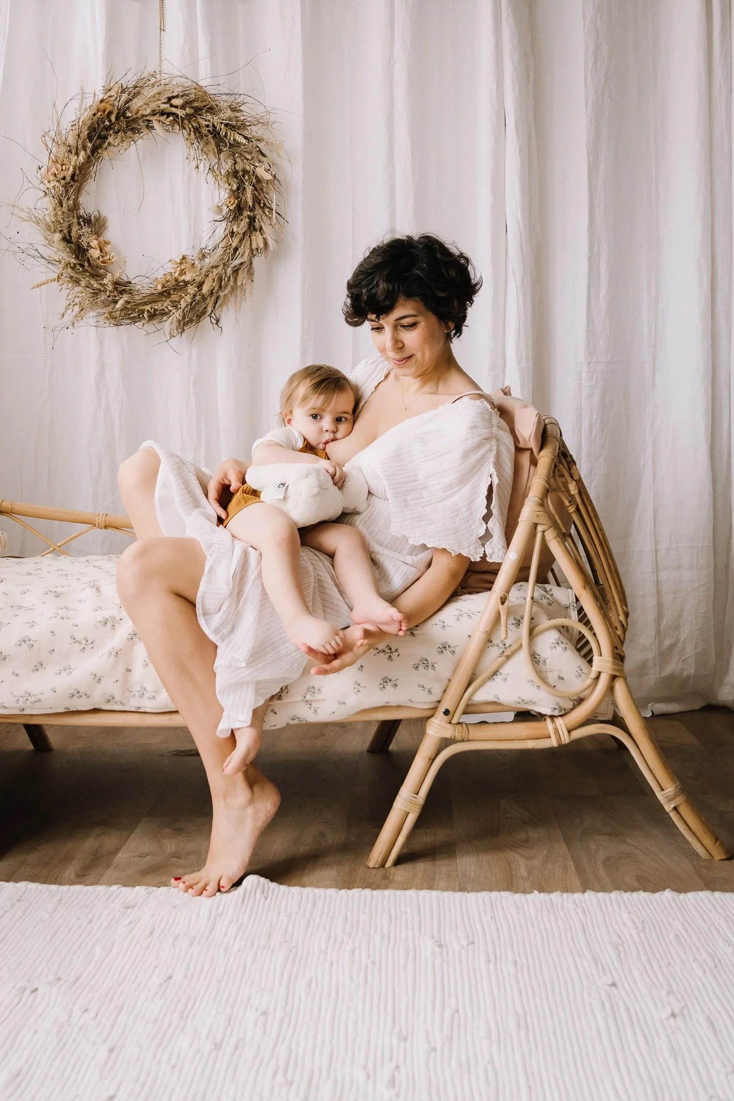 A woman holding a young child on her lap, sitting on a wicker bench in a cozy room with white curtains and a floral cushion, decorated with a wreath on the wall.