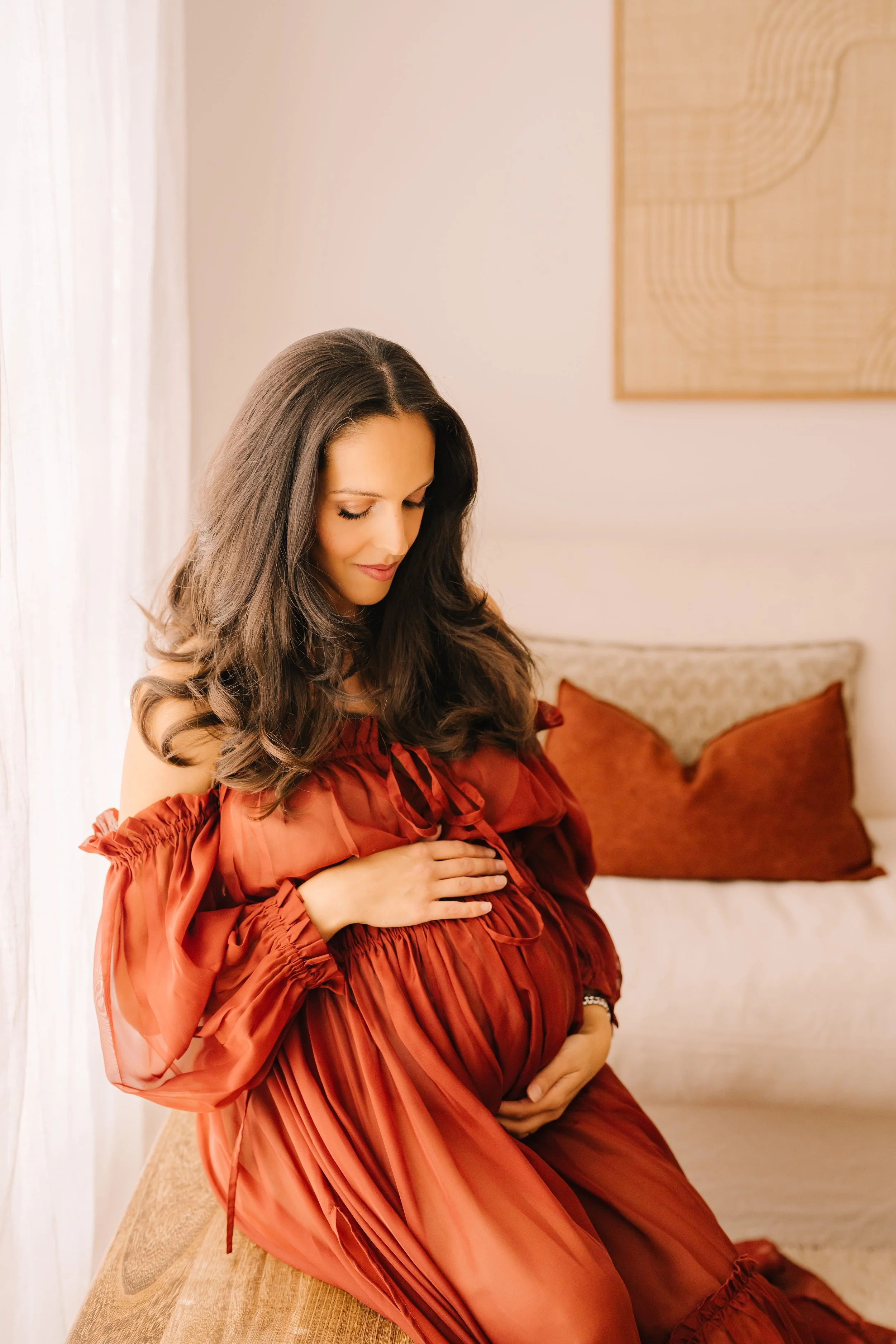 Pregnant woman in a rust-colored dress sitting on a bench, gently cradling her belly and looking down with a smile.