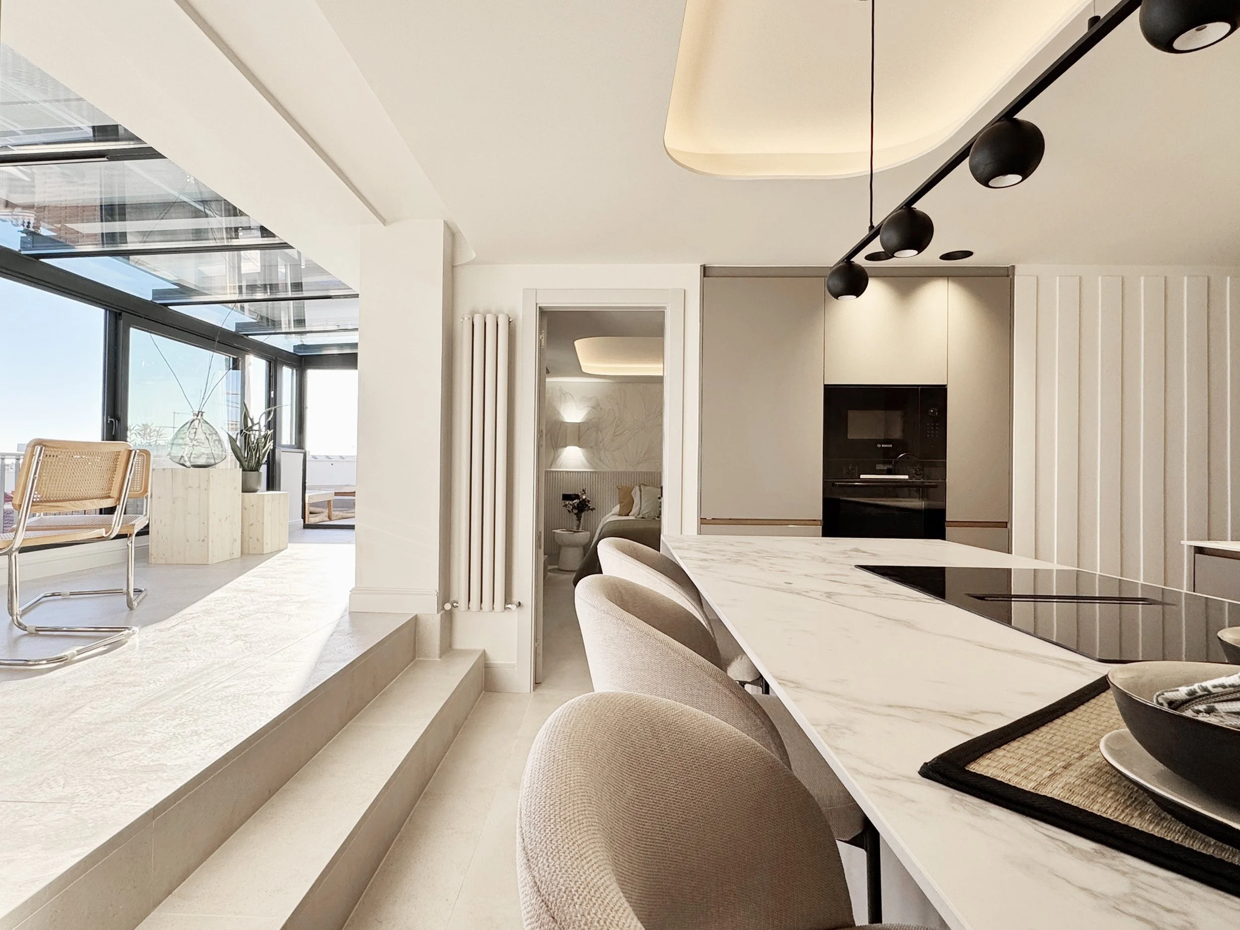 Cocina moderna con isla de mármol y sillas beige, con luz natural que entra por ventanas grandes, y una vista al balcón con plantas decorativas.