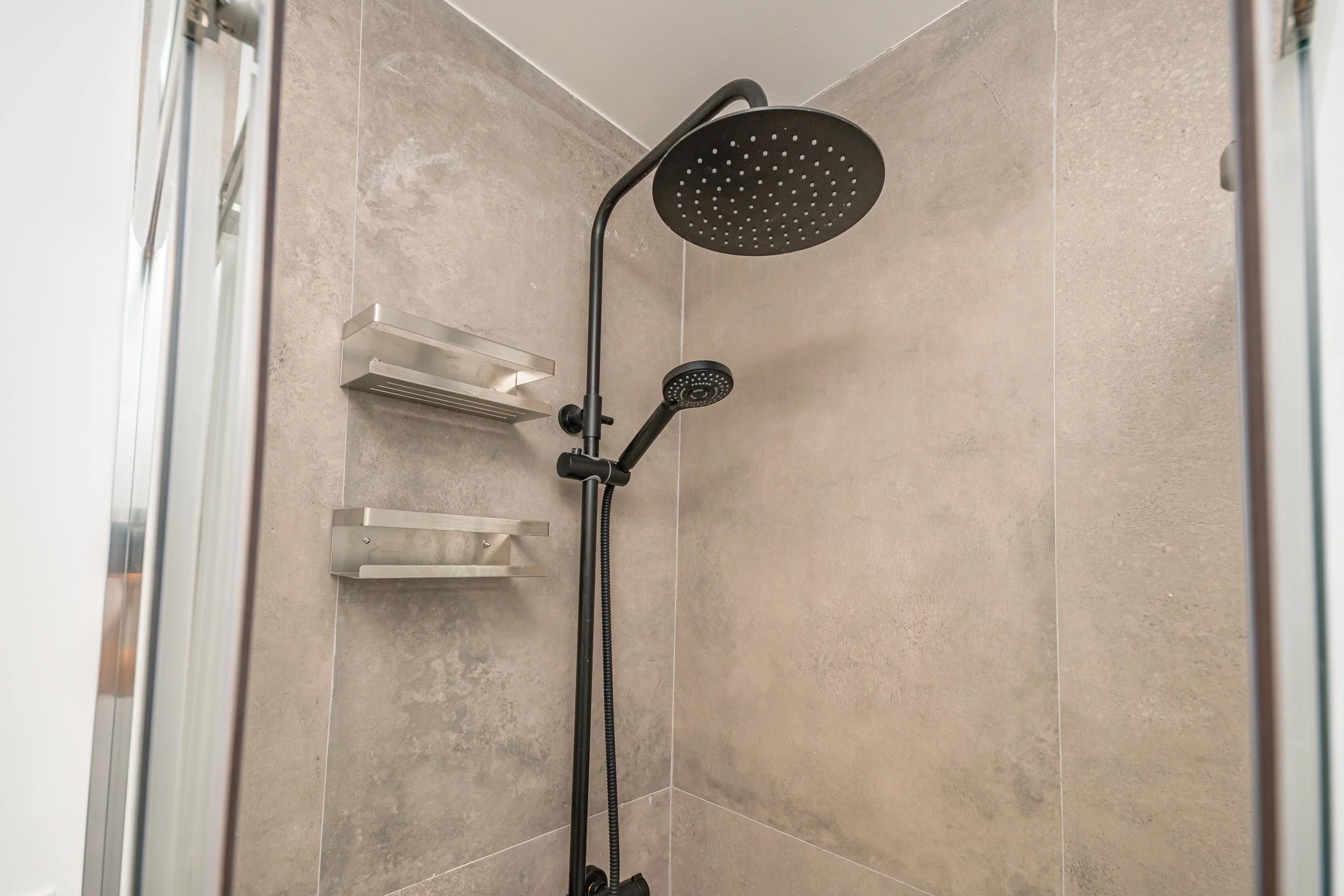 Regadera de ducha negra con cabezal grande y manguera en una pared de azulejos beige.