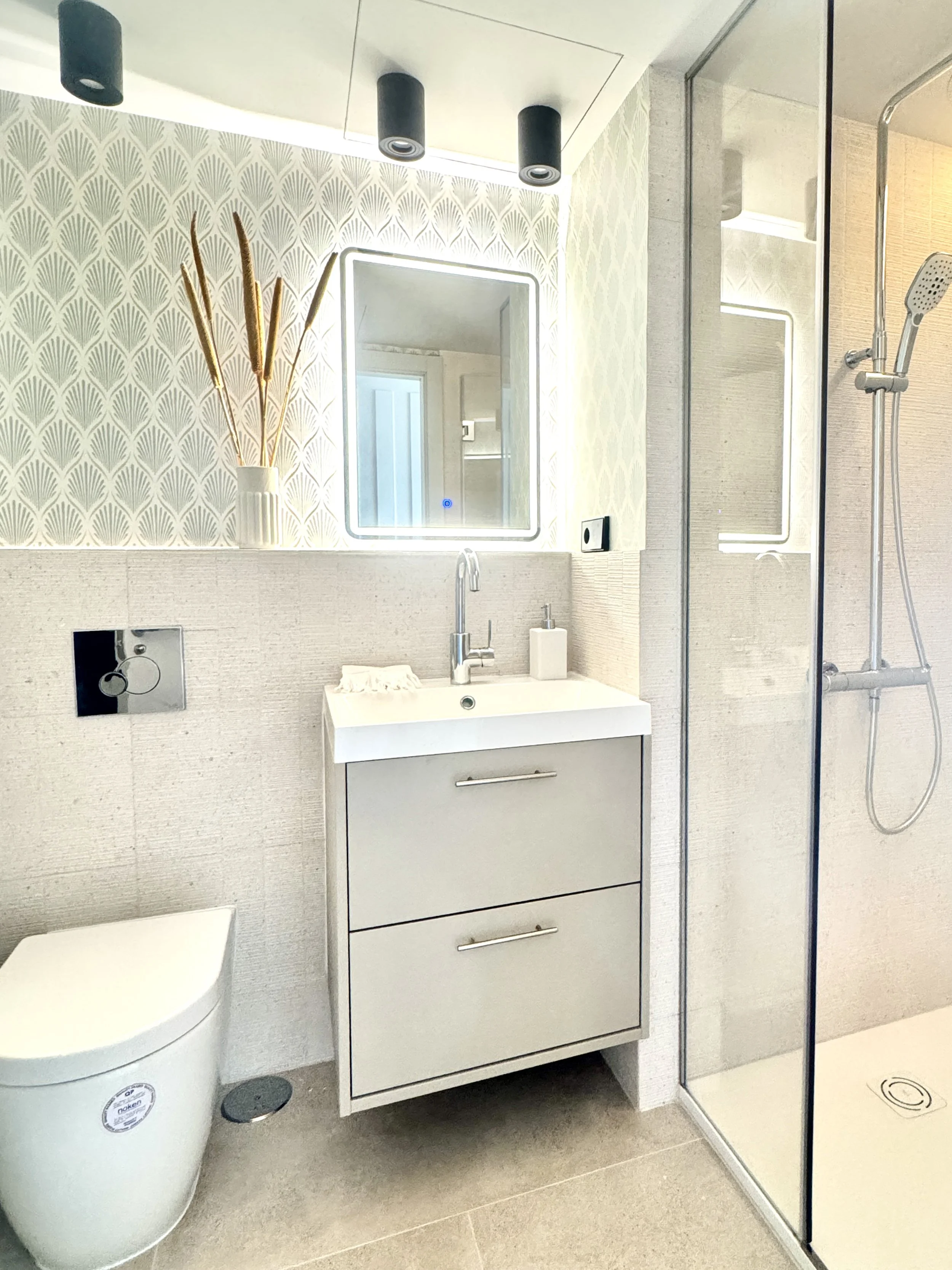 Baño moderno con espejo con iluminación, lavabo con dos cajones, ducha con mampara, taza sanitaria y decoraciones en la pared.