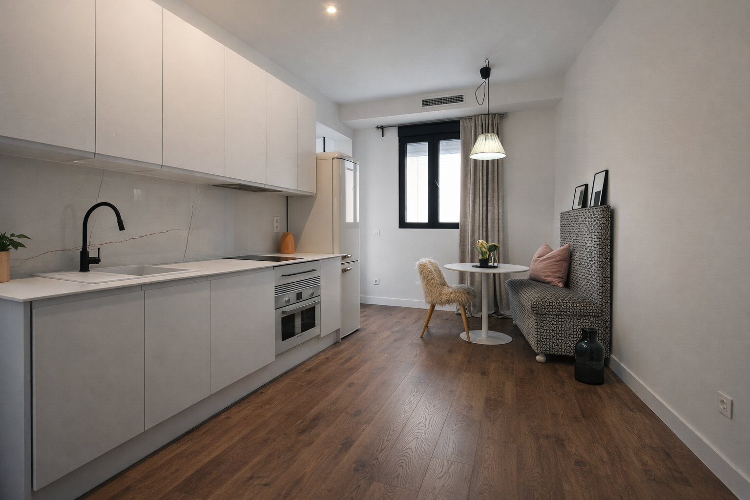 Sala pequeña con cocina blanca y mesa con silla de peluche y sofá con cojín rosado, en una habitación con suelo de madera y ventanas con cortinas beige.
