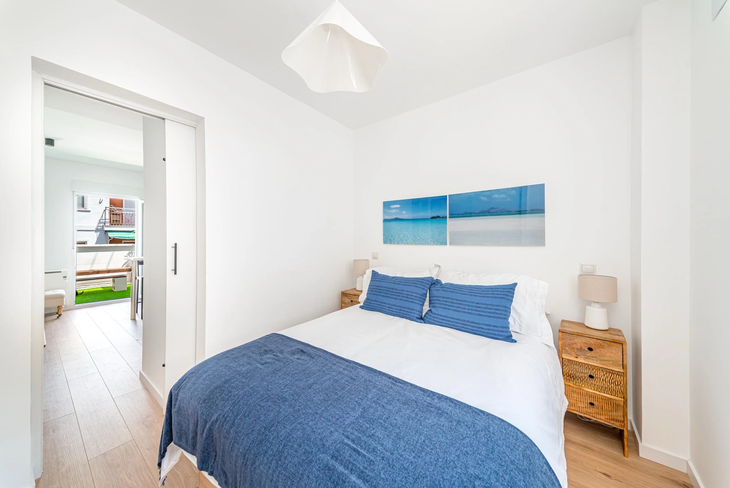 Dormitorio con paredes blancas, cama con sábanas blancas, y almohadas azules, alféizar con arte de paisaje marino