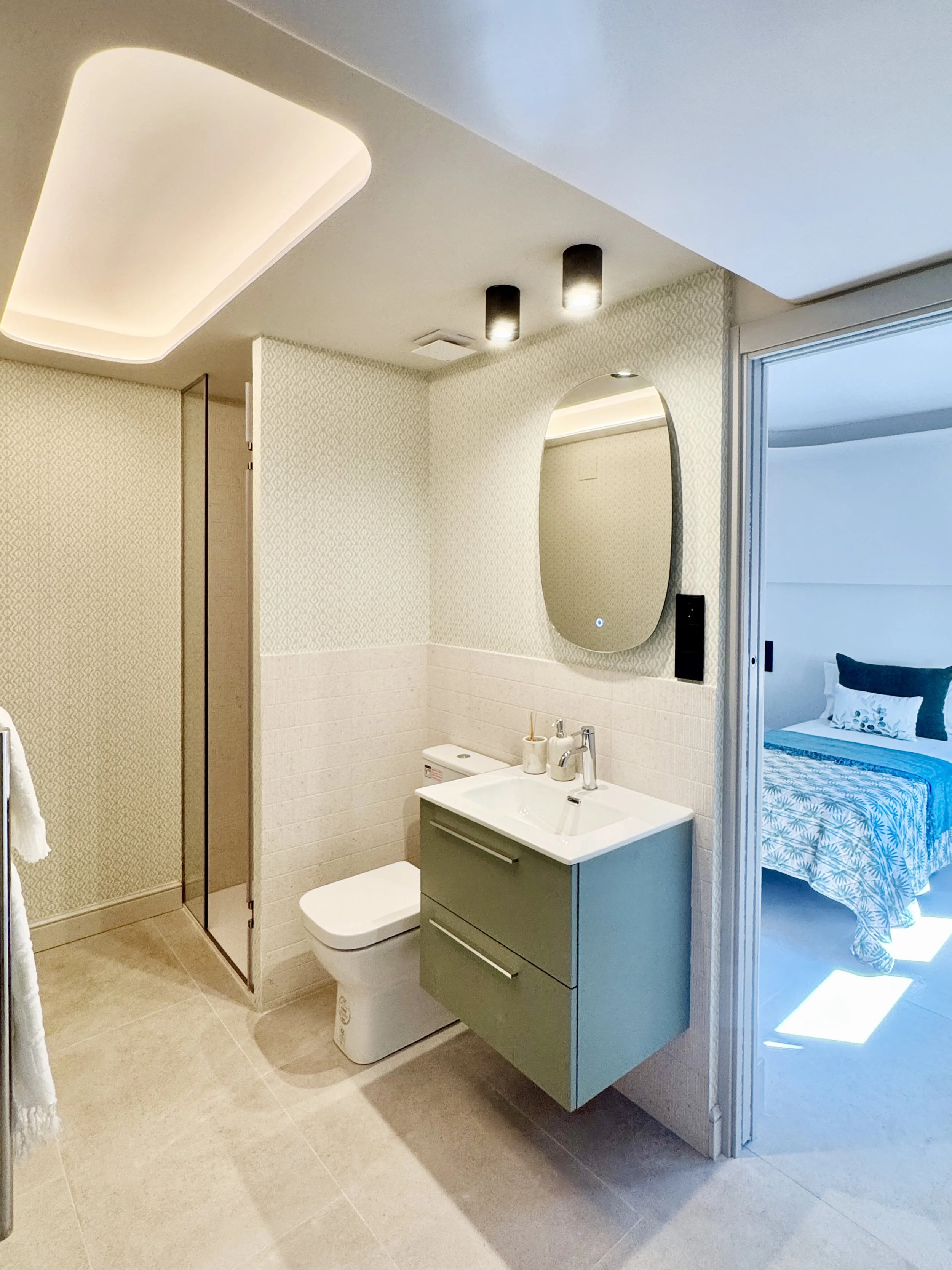 Baño moderno con espejo ovalado, lavabo con mueble verde, inodoro y lavandería, conectado a un dormitorio con cama y ropa de cama azul.