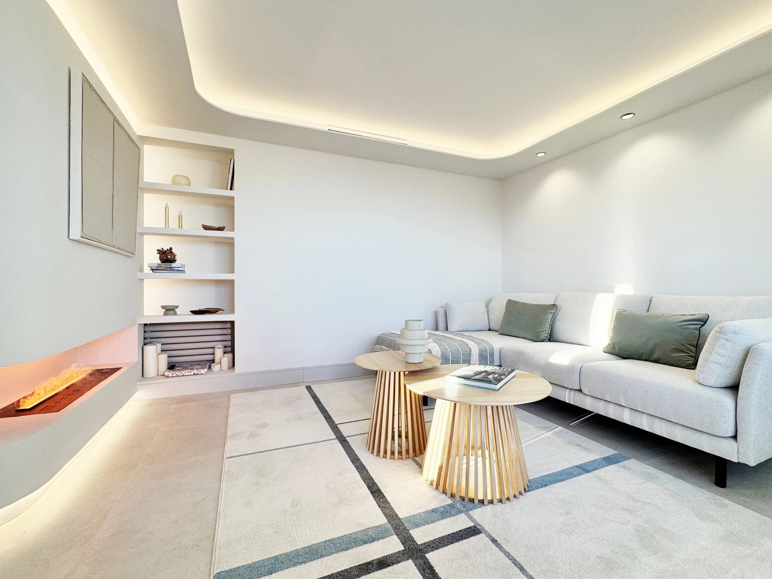 Sala moderna con sofá gris, mesas de madera, estantería blanca decorada con objetos, chimenea con luces cálidas, y alfombra clara con líneas negras y azules.